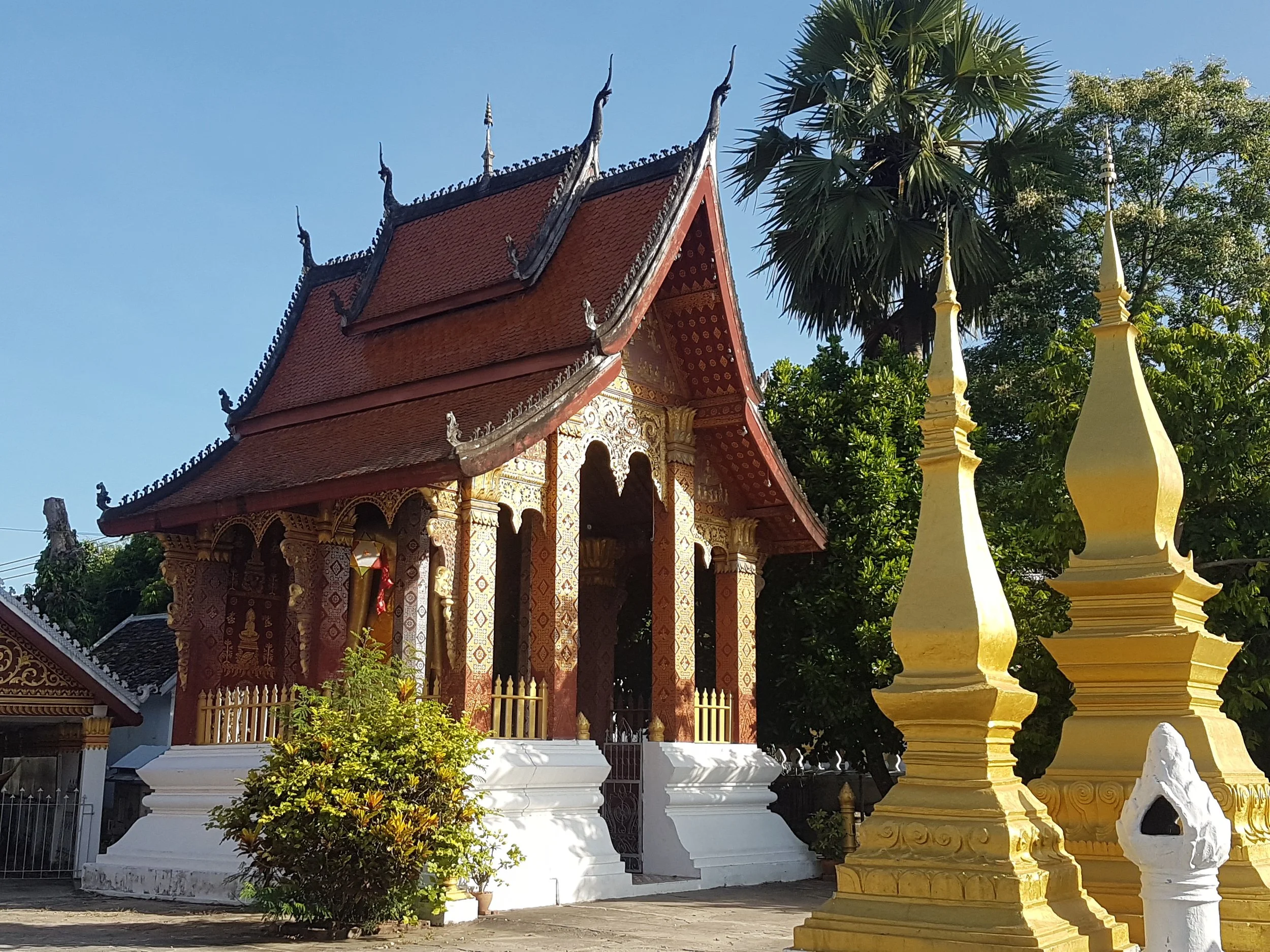 Temple Bouddhiste traditionnel avec toit en pente, décorations dorées, entouré d'arbres et de stupas dorés dans un cadre extérieur ensoleillé.