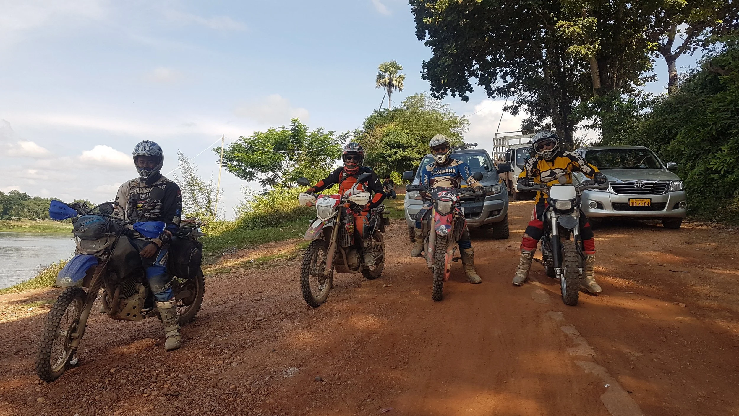 Cinq motards portant un équipement de protection, debout ou assis sur leurs motos tout-terrain, devant des voitures garées dans un environnement naturel avec des arbres et un ciel partiellement nuageux.