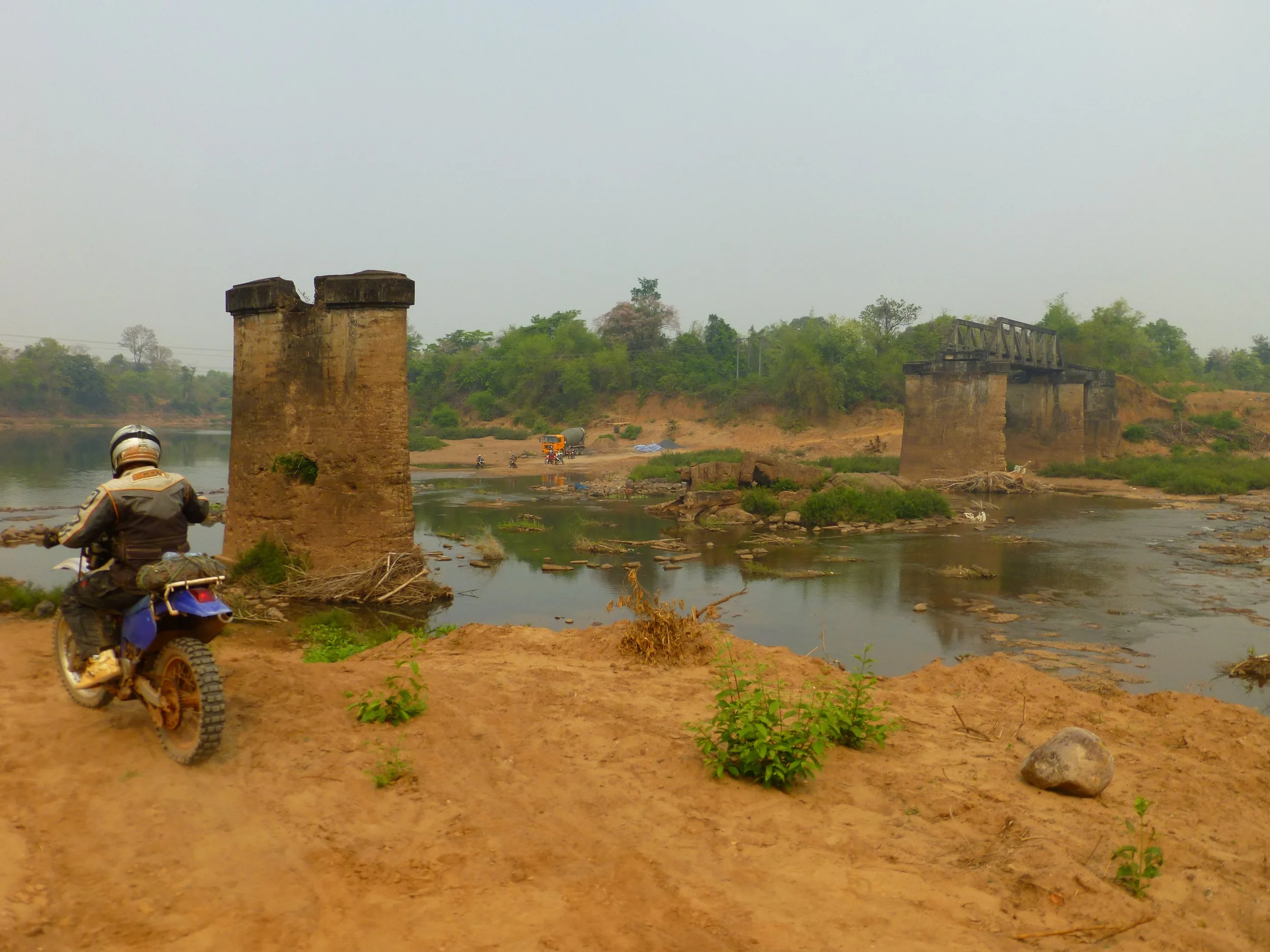 Un pont en ruine au dessus d'une rivière avec un pilote de moto en premier plan, mais son visage n'est pas visible.
