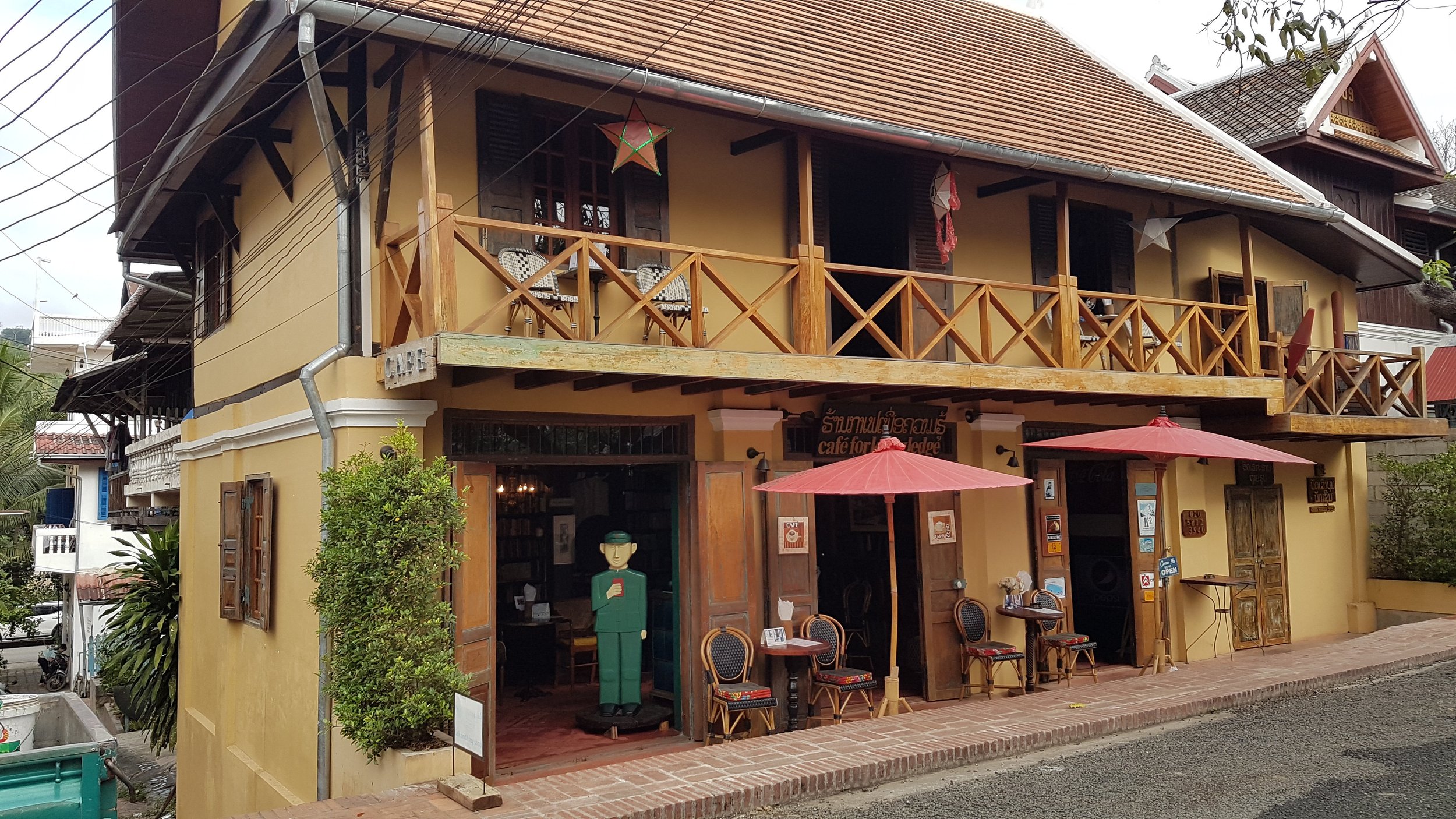 Un bâtiment de style asiatique avec un balcon en bois, une enseigne de café, des parasols rouges et des meubles extérieurs, entouré de végétation.