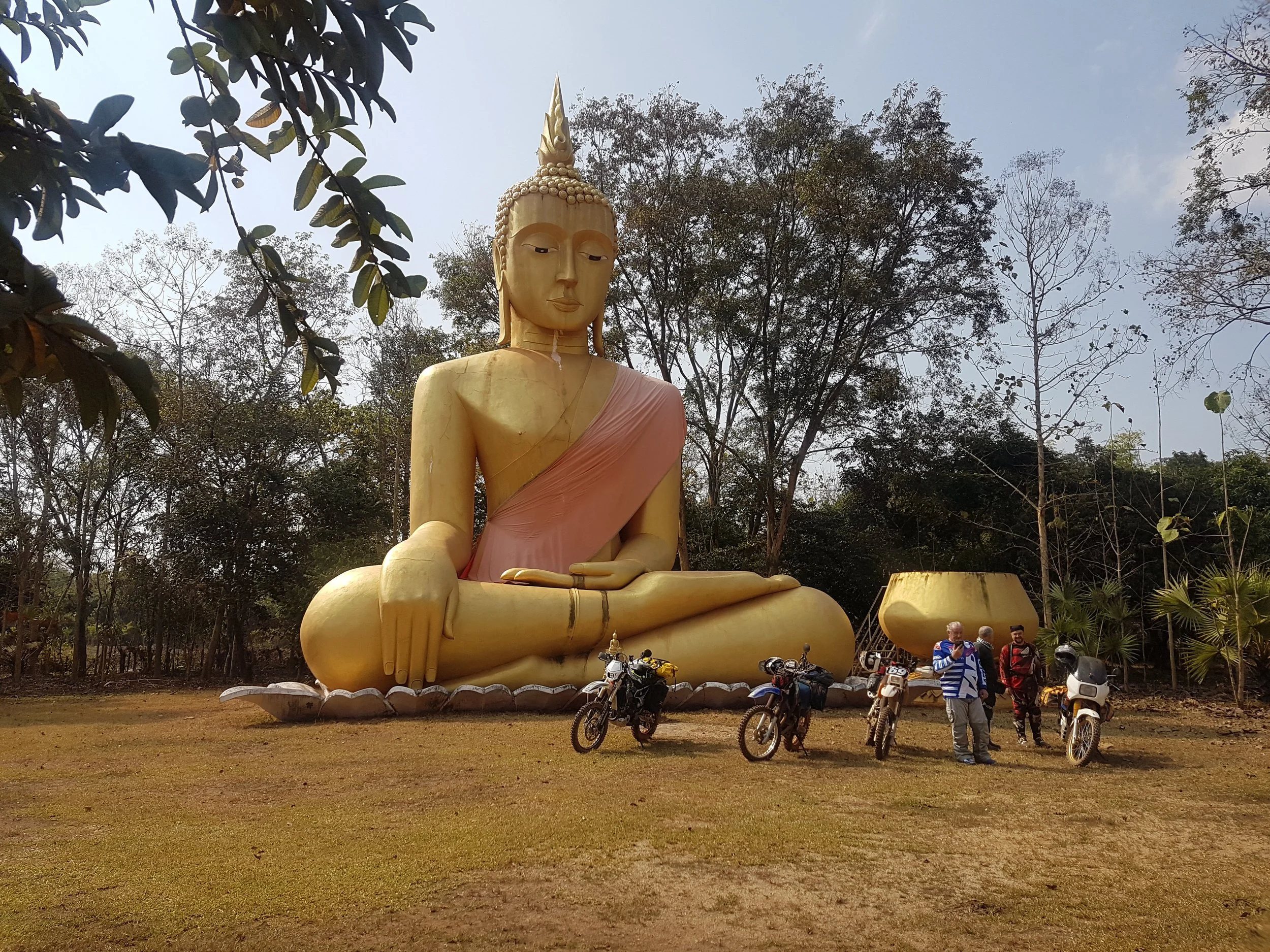 Grande statue dorée de Bouddha assis dans un environnement naturel avec des arbres, des personnes et des motos en premier plan.