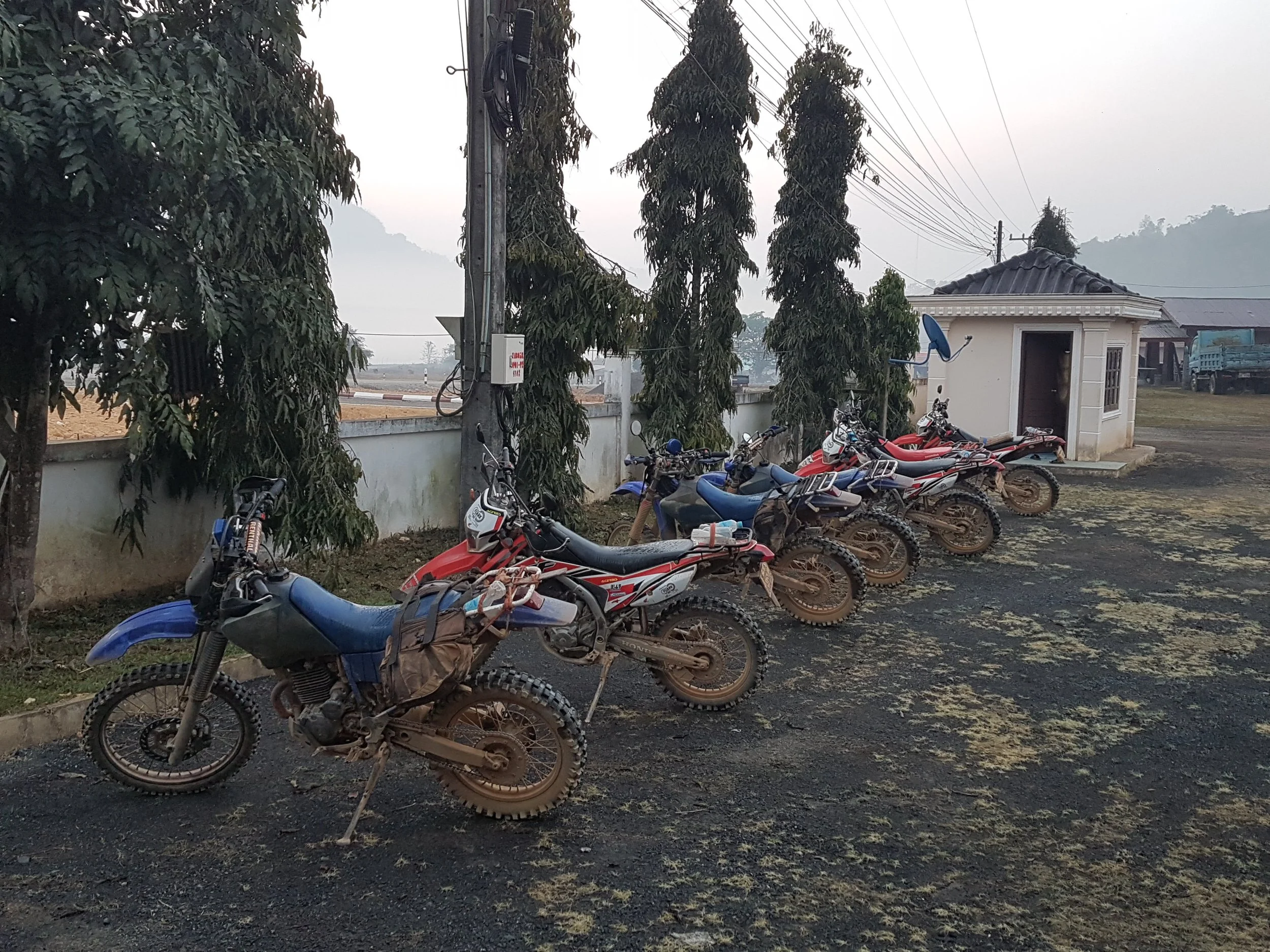 Une rangée de motos rouges et bleues garées sur un sol sombre, avec un petit bâtiment blanc avec un toit en tuiles gris à droite, des arbres en arrière-plan et des câbles électriques dans le ciel brumeux.