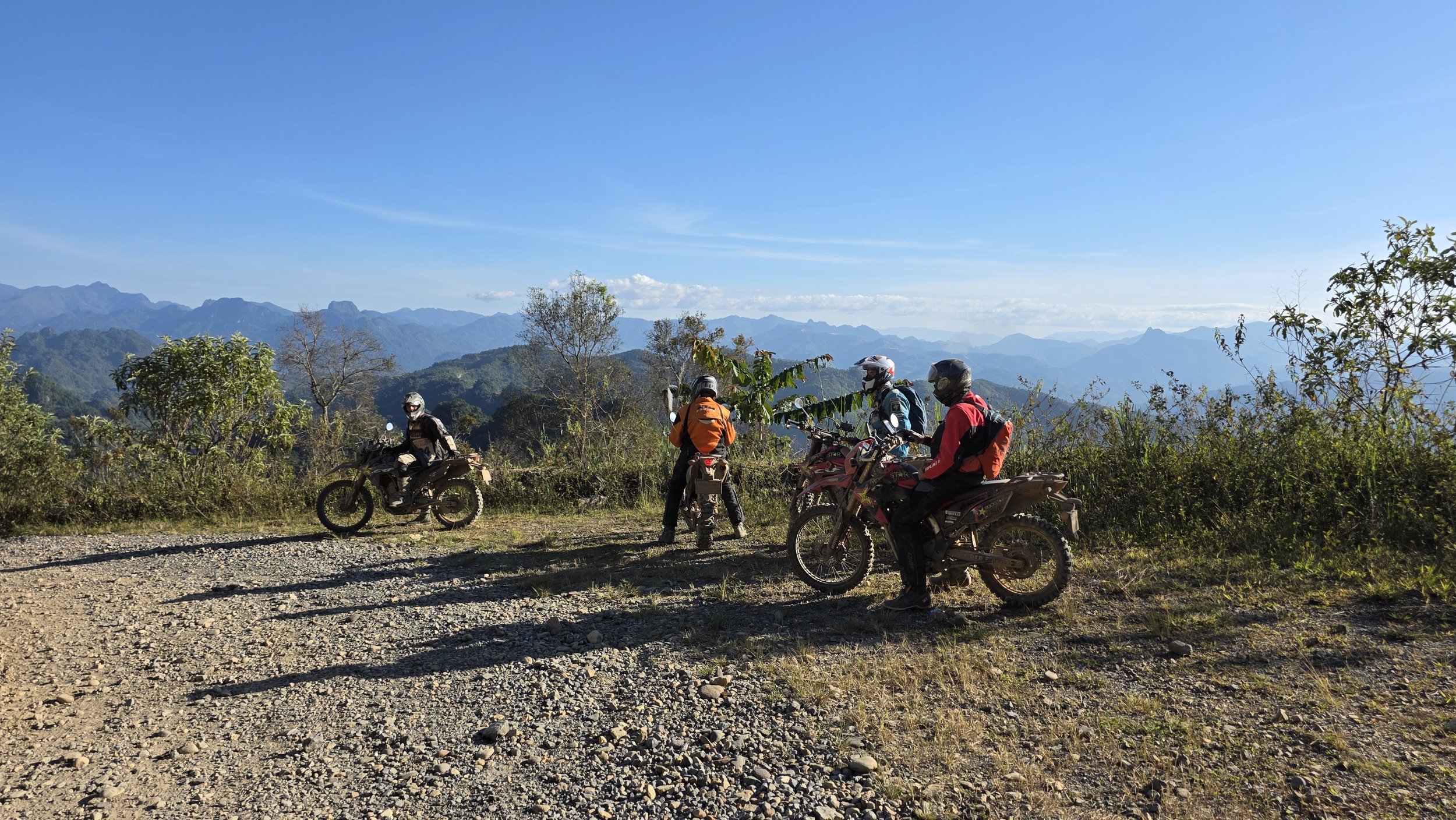 Groupe de motards avec casques et équipements sur une route de montagne, paysage de montagnes en arrière-plan, ciel clair.