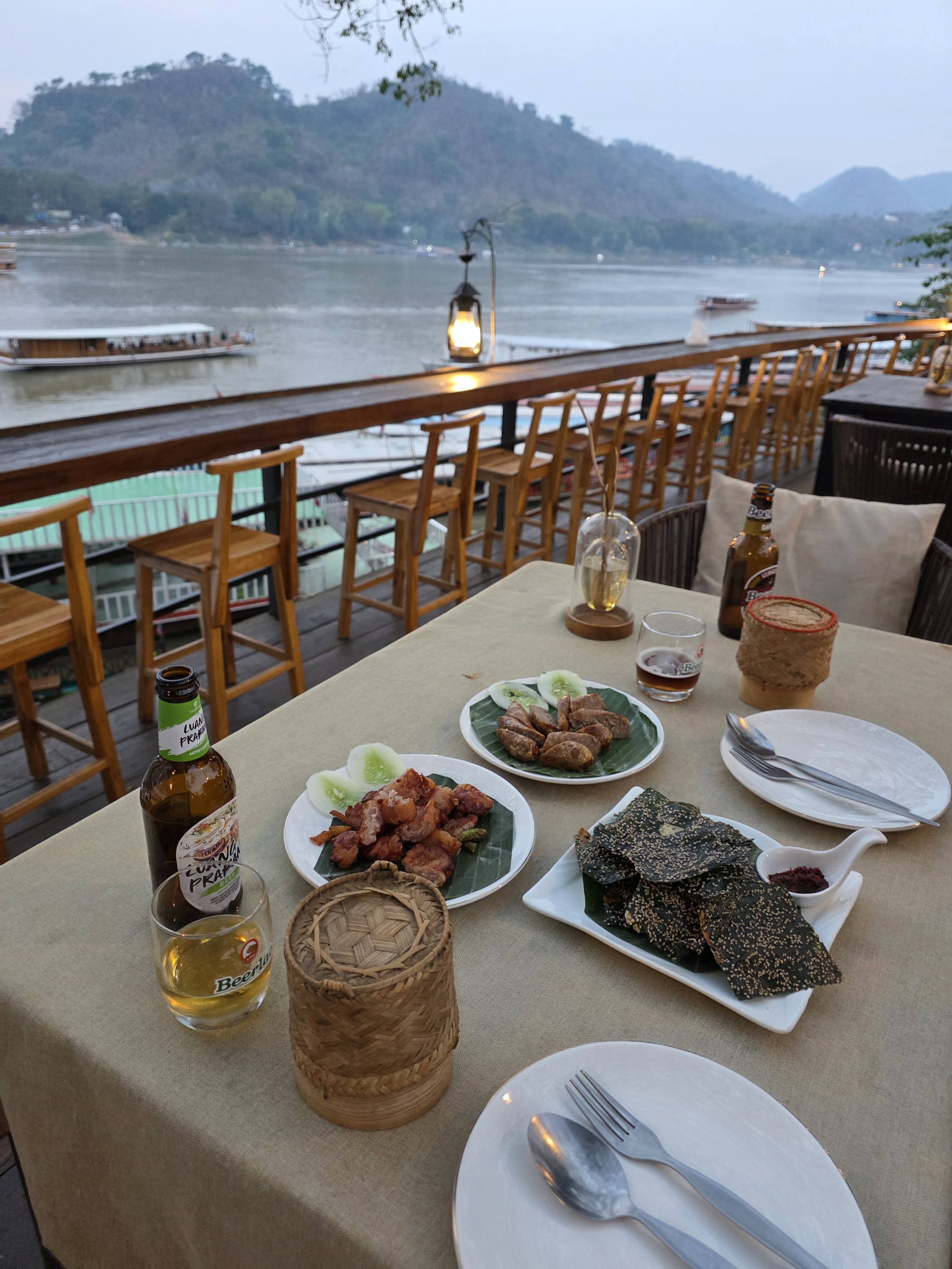 Repas thaïlandais avec des plats de nems, viandes et légumes, bière et vue sur le fleuve avec des bateaux et des montagnes en arrière-plan.