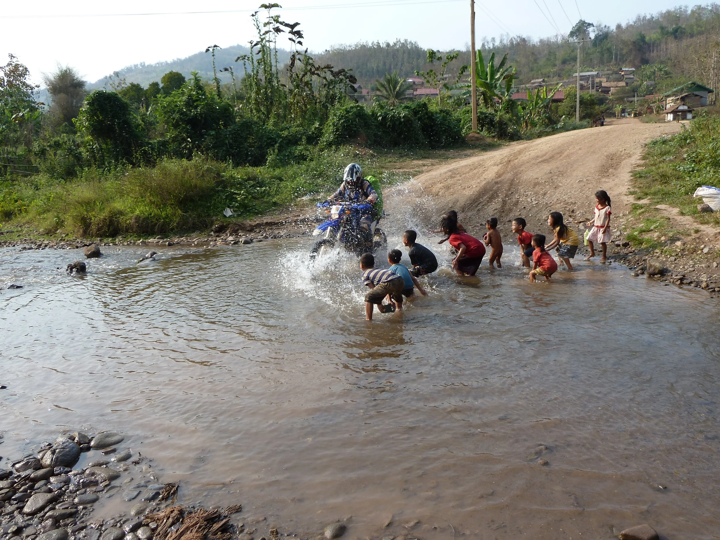 Un enfant en casque de moto traverse une rivière à moto, entouré d'enfants et d'adultes sur la berge dans un village rural en hauteur avec des maisons en bois et des arbres luxuriants.
