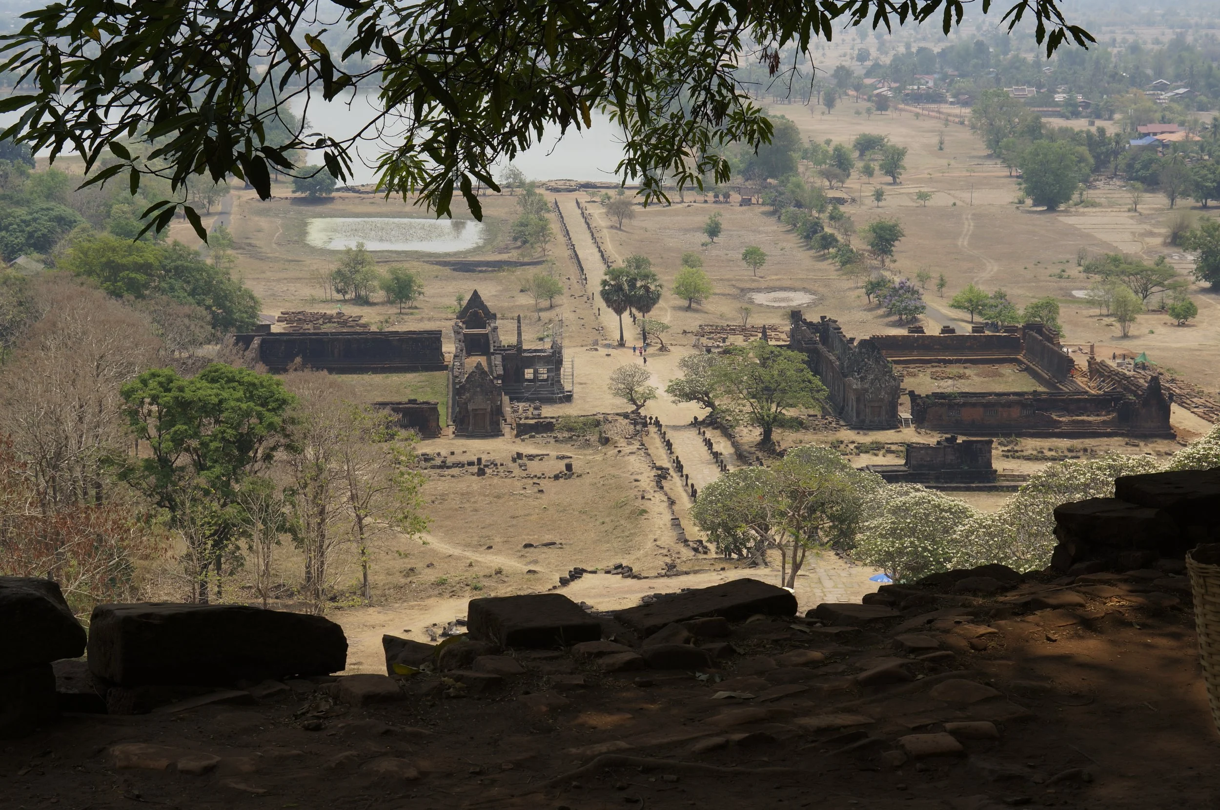 Vue d'un site archéologique avec des ruines anciennes entouré de végétation et de champs agricoles, prise depuis une grotte ou un endroit abrité.