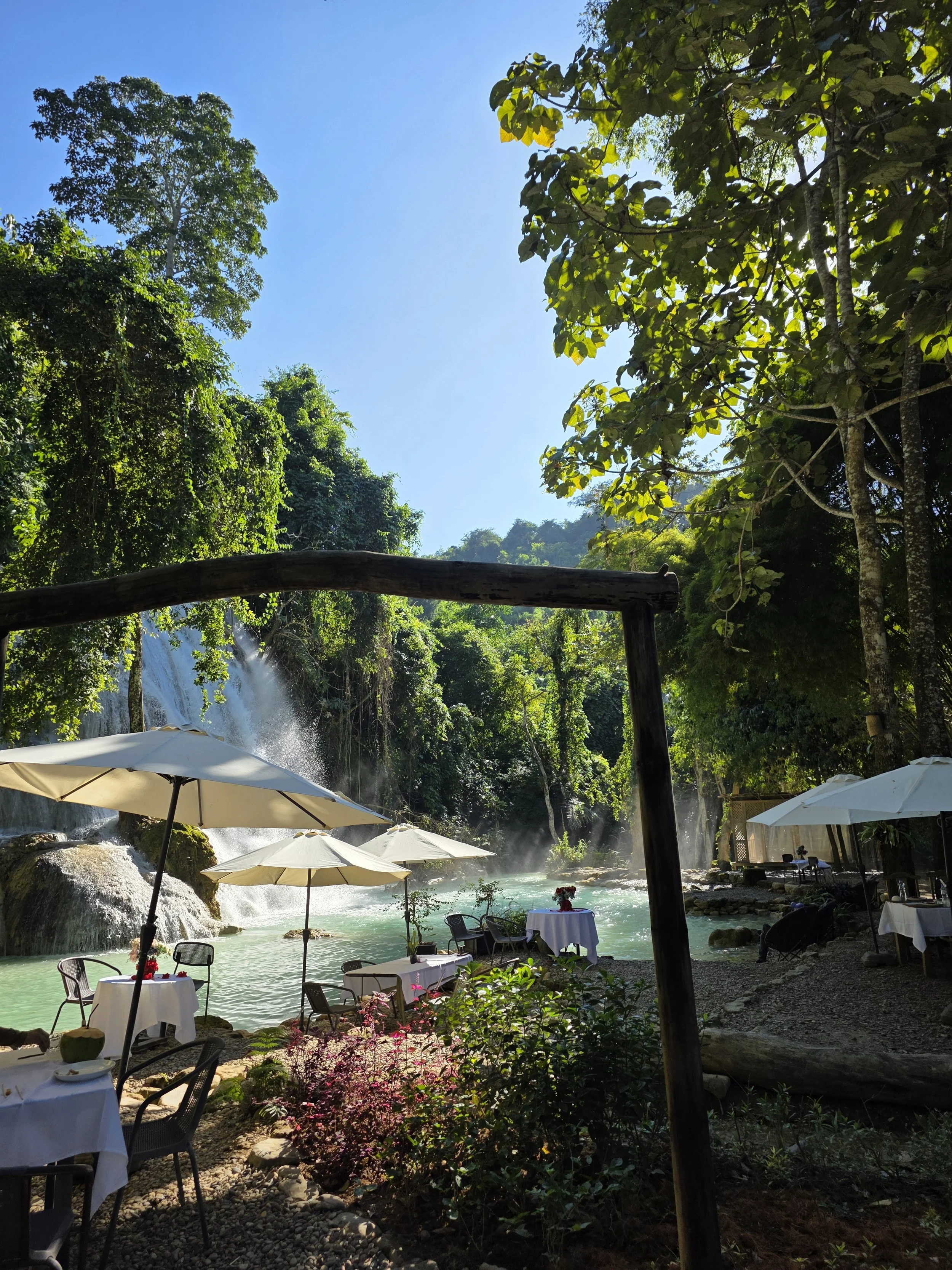 Un restaurant en plein air au bord d'une rivière ou d'une chute d'eau, entouré de forêts, avec des tables, des parasols et un décor naturel assemblé sous un soleil clair.