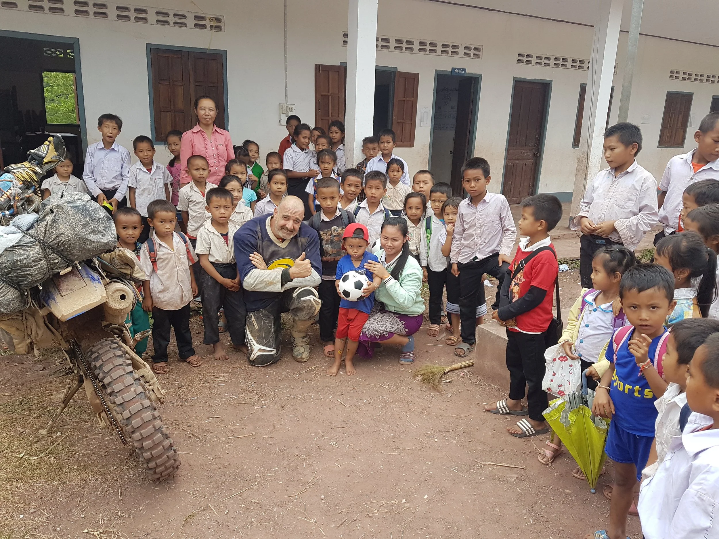 groupe d'enfants de différentes ethnies avec deux adultes devant une école en milieu rural, certains portant des sacs à dos, un garçon tenant un ballon de football, et un motocycle en premier plan.