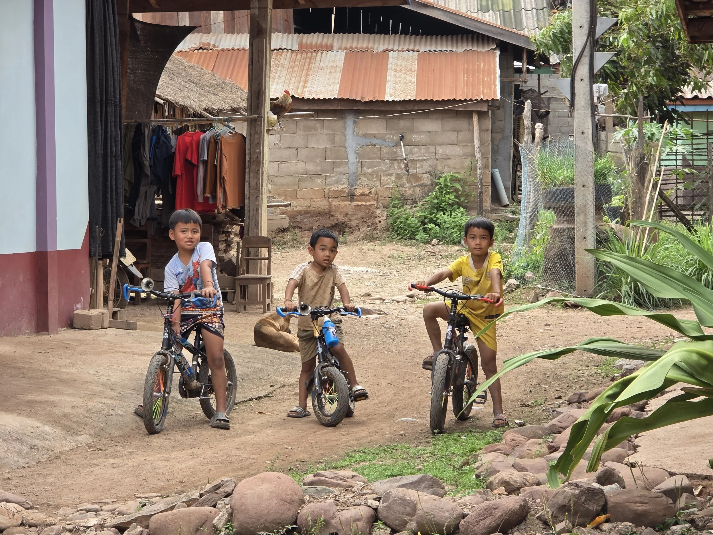 Trois garçons dans un village rural, avec des vélos, à côté d'une maison en briques avec un toit en tôle ondulée, et un rocher, dans une zone végétale