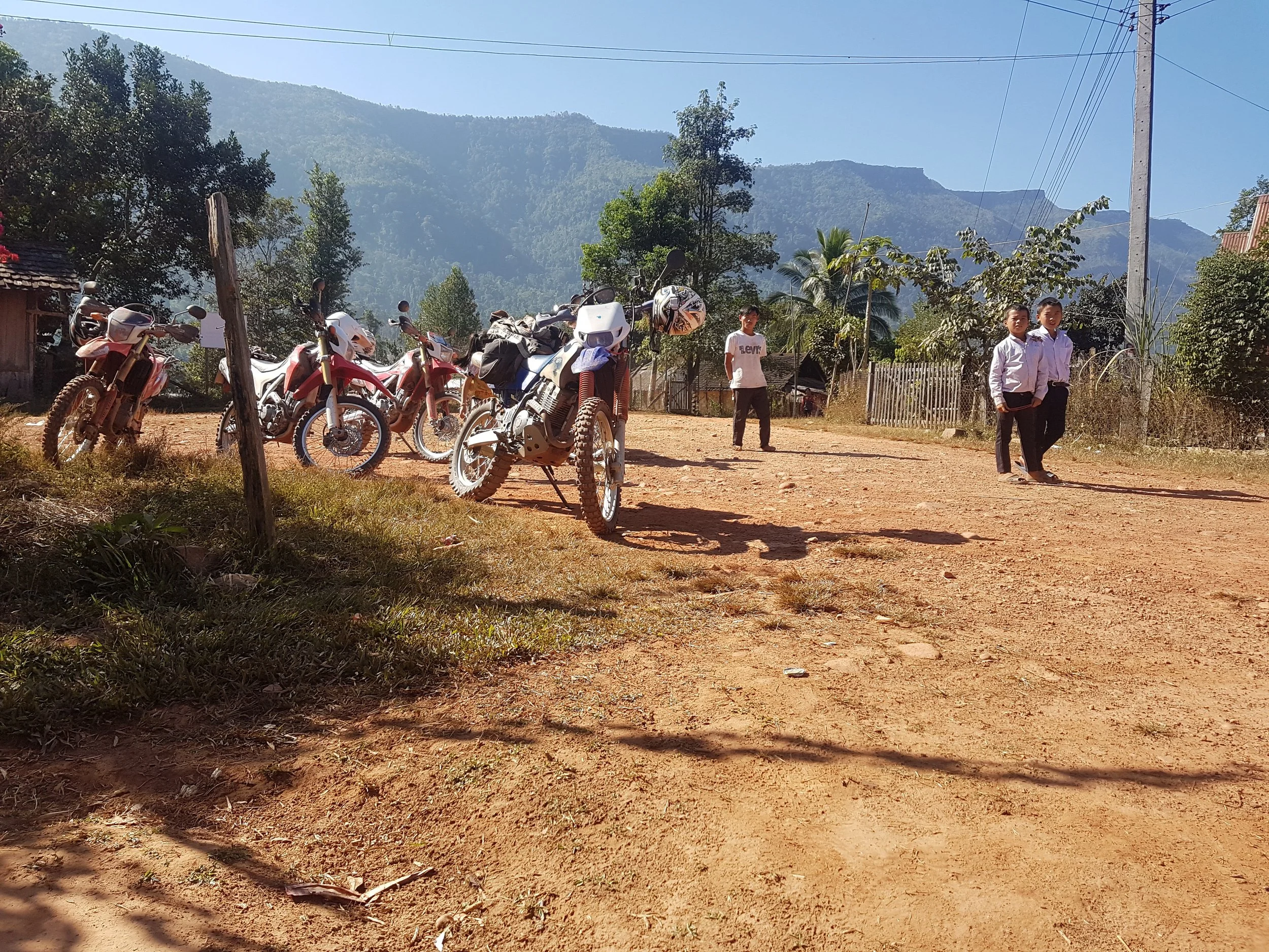 L'image montre un terrain en terre rouge avec plusieurs motos garées, deux enfants en uniformes scolaires marchant, entourés d'arbres et de montagnes en arrière-plan, sous un ciel clair.