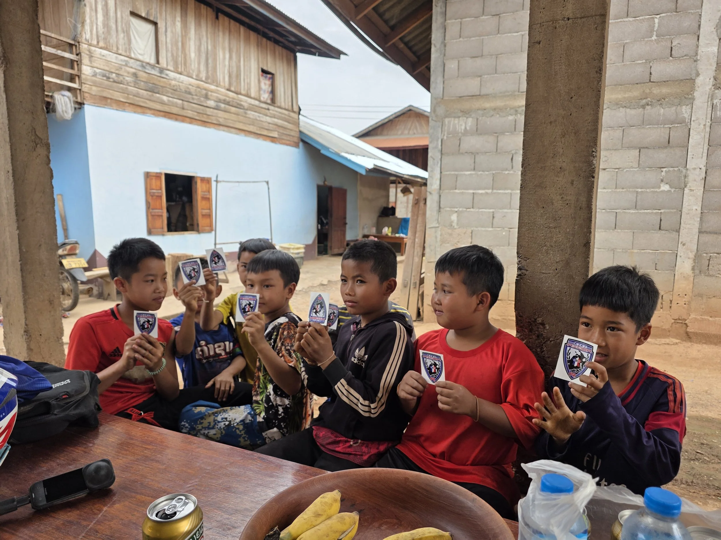 Groupe d'enfants souriants assis à une table en plein air, tenant des badges ou des autocollants avec un logo de sport ou d'équipe.