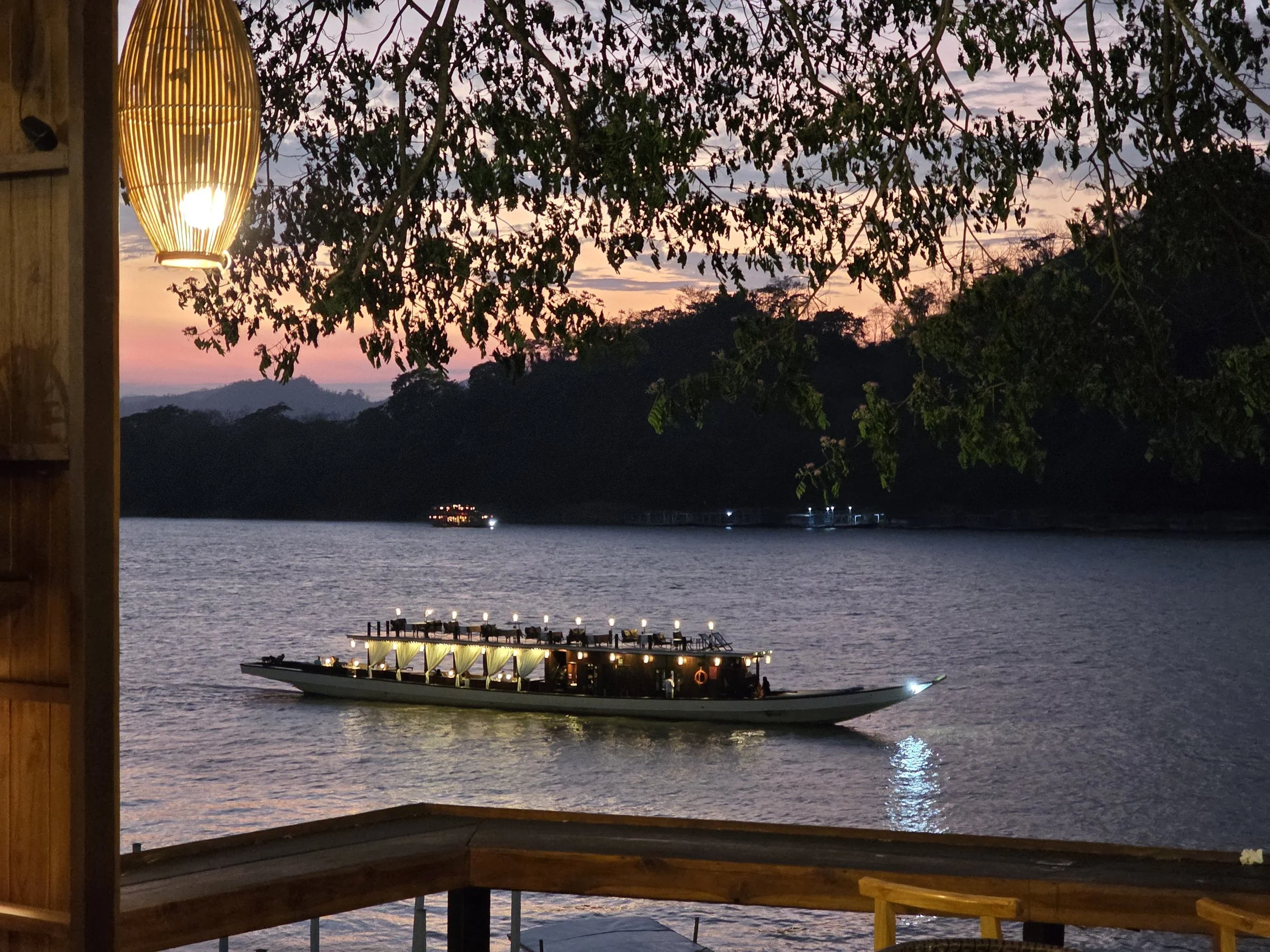 Un bateau de croisière éclairé navigue sur une rivière au coucher du soleil, avec des collines en arrière-plan et des arbres en premier plan.