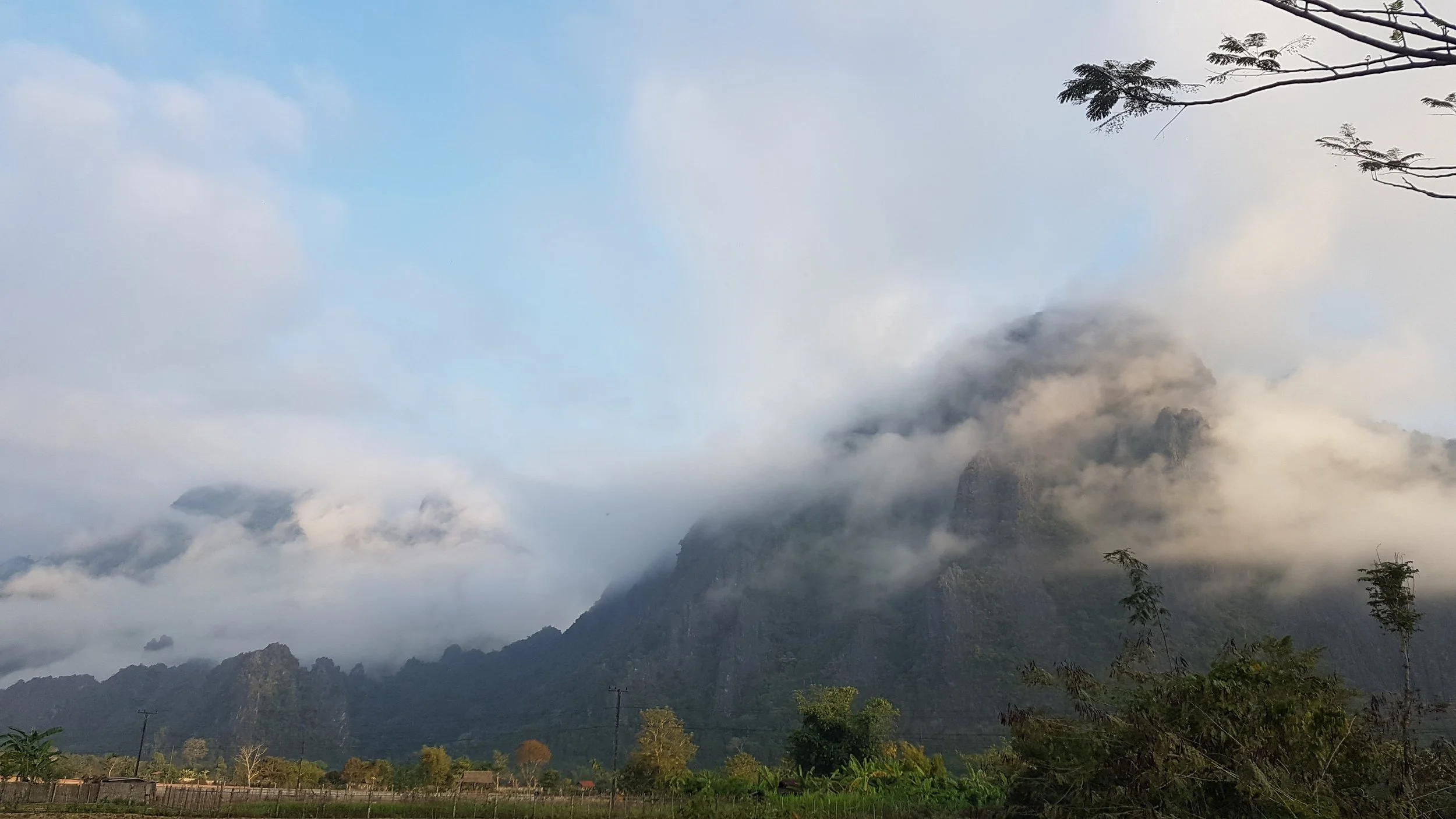 Montagne enveloppée de nuages, arbres en premier plan, ciel partiellement bleu.