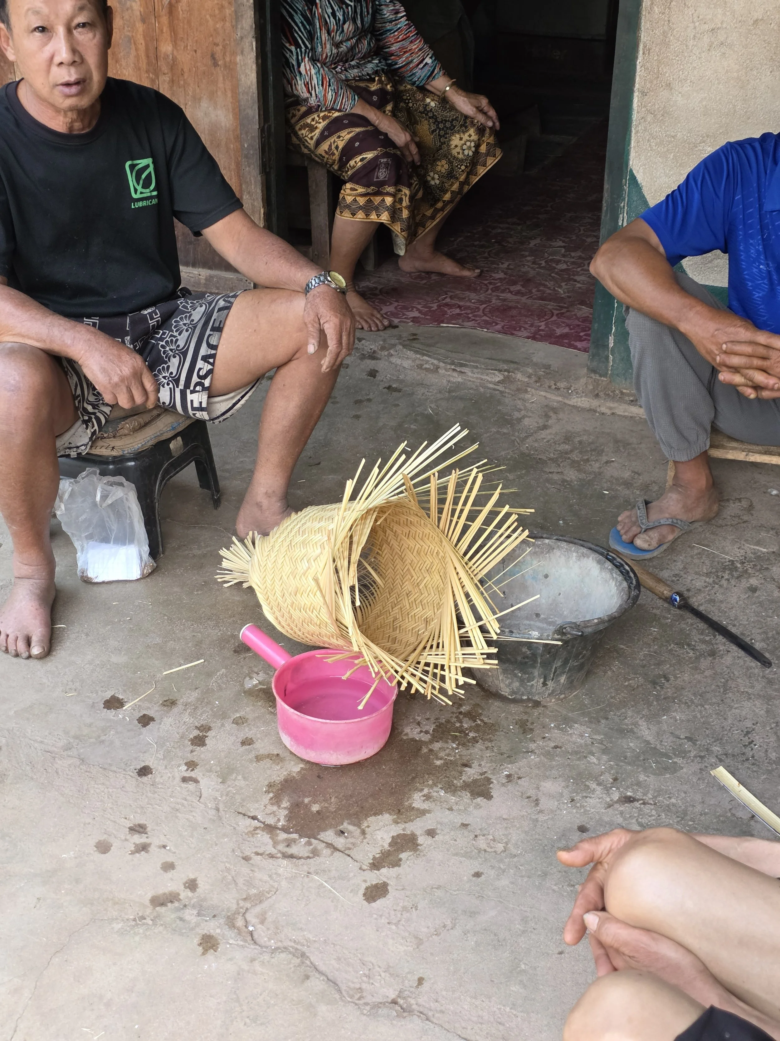 Un groupe de personnes assises à l'extérieur, avec un panier de paille et un seau rose sur le sol. Il semble que des activités artisanales ou culinaires soient en cours.