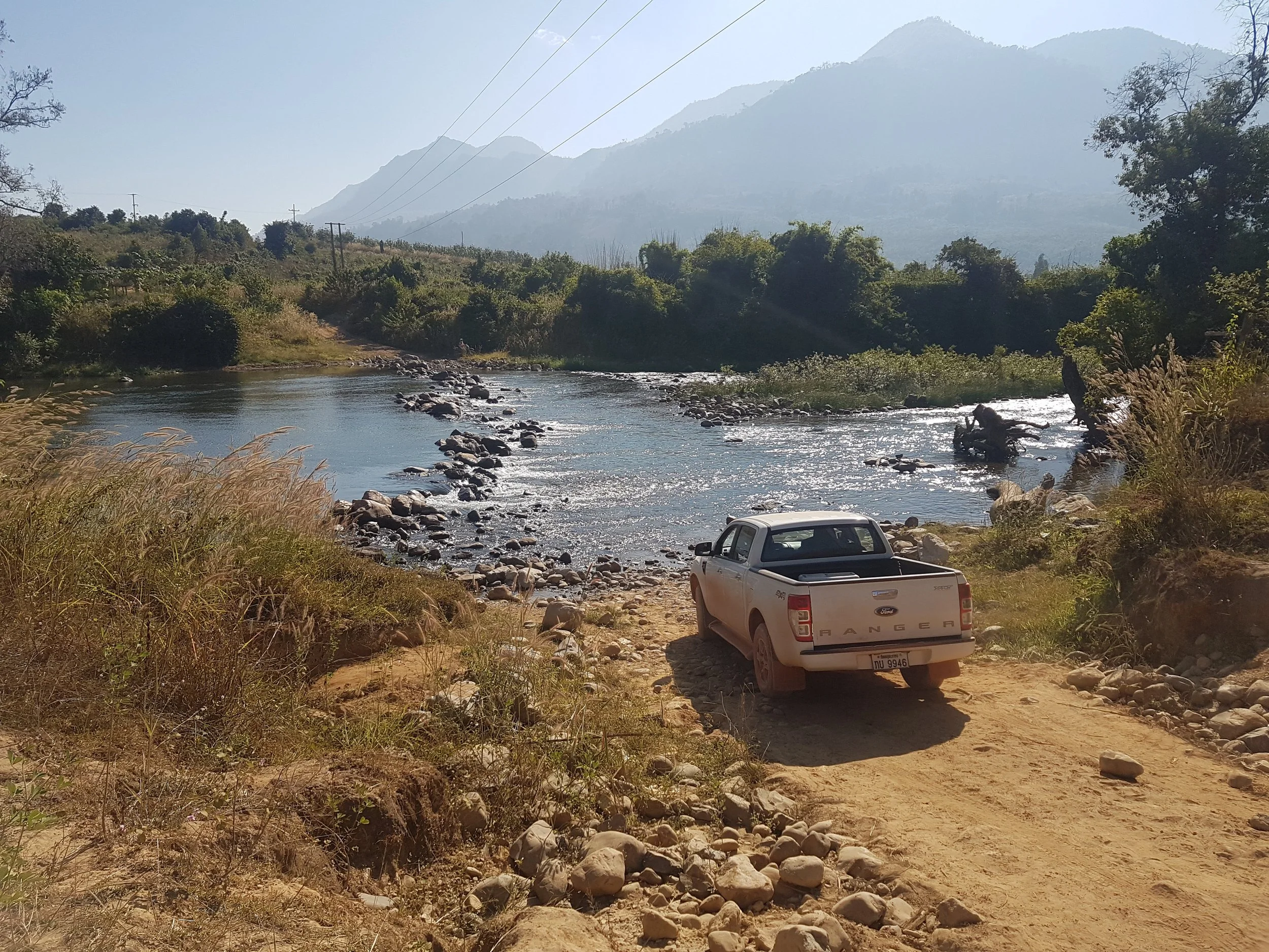 Une rivière traversant un paysage naturel avec des montagnes en arrière-plan, des arbres et de la verdure, un véhicule blanc garé sur un chemin de terre.