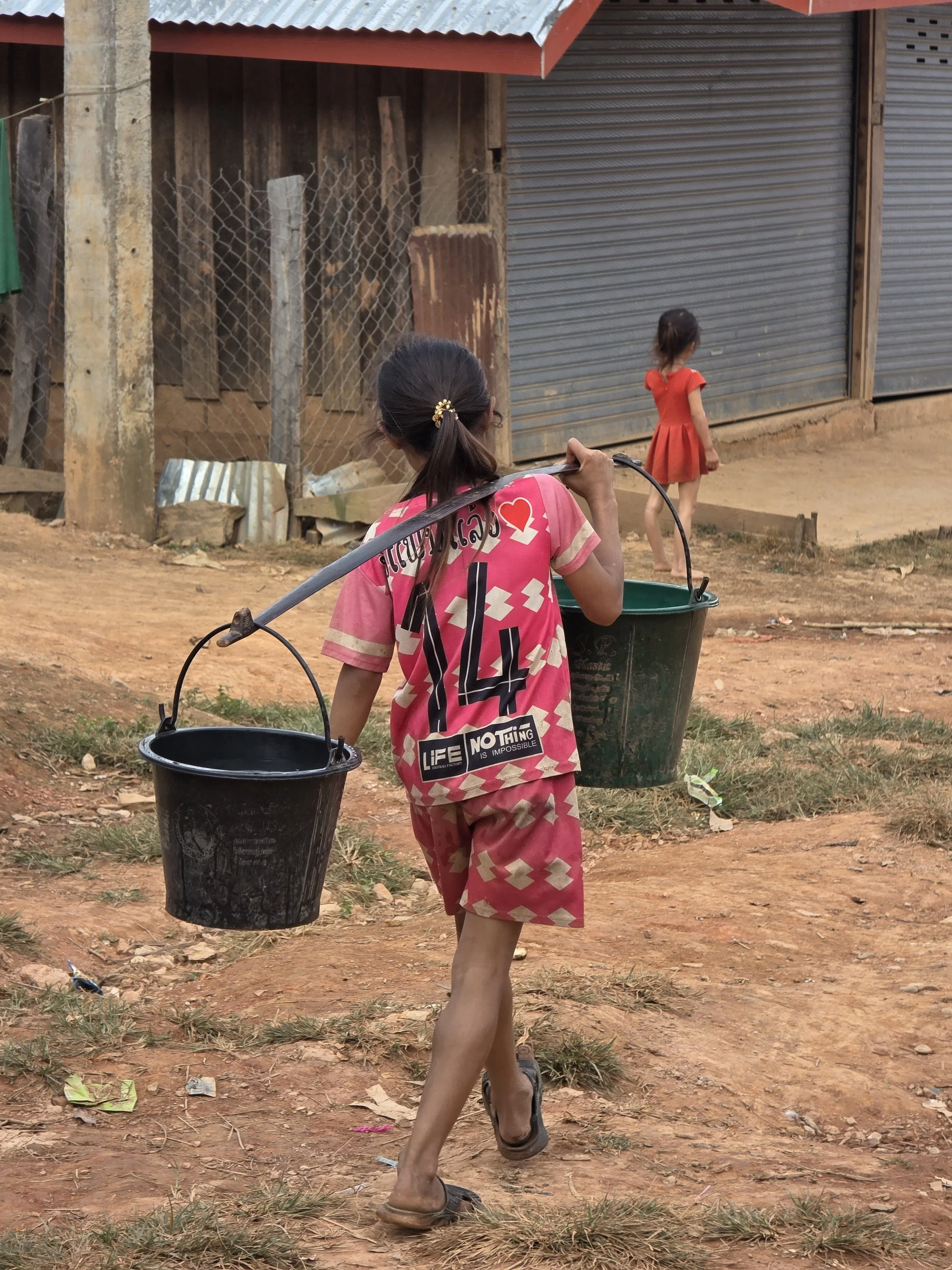 Une jeune fille marchant sur un chemin en terre avec deux seaux suspendus à une tige en métal. Une autre enfant, portant une robe orange, marche devant une boutique avec des volets métalliques fermés. La scène semble se dérouler dans un environnement
