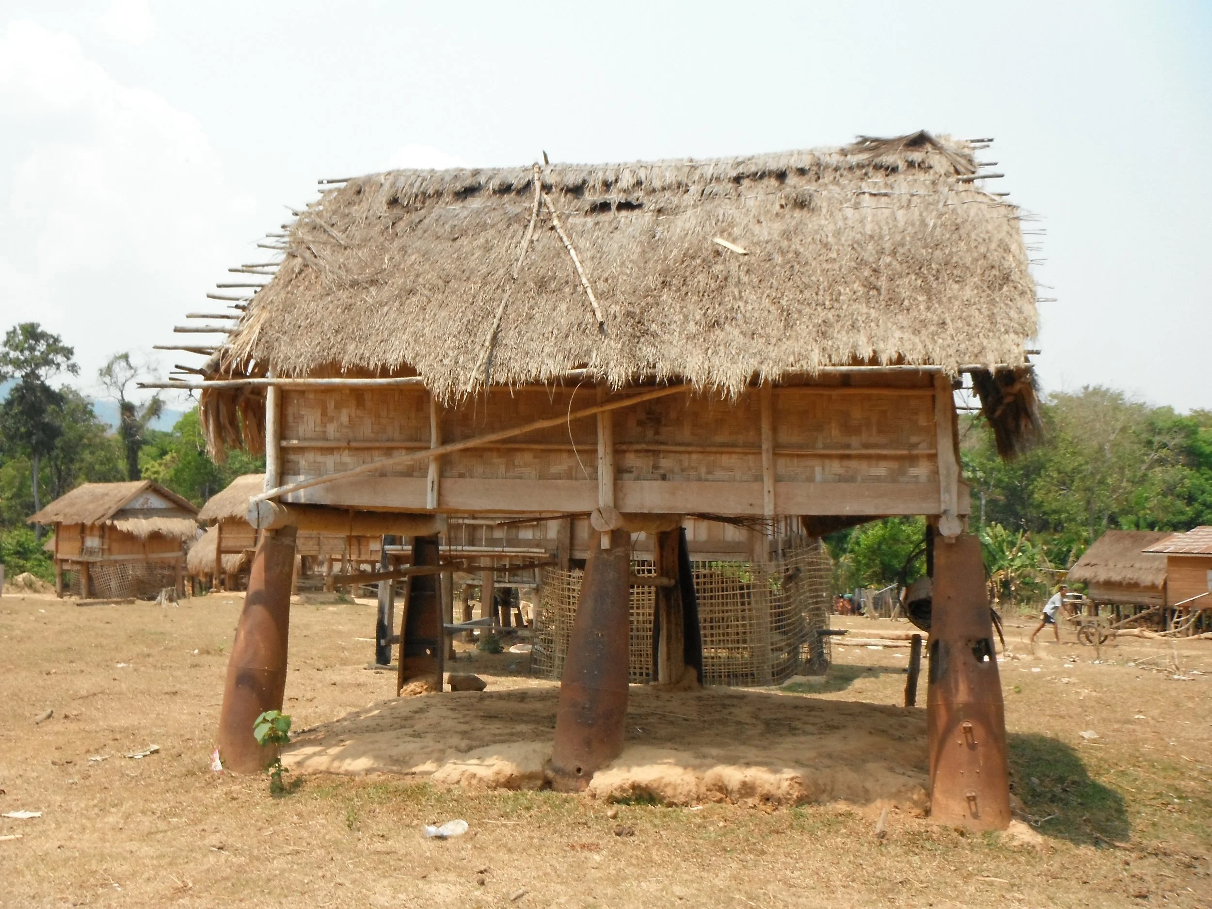 Maison sur pilotis avec toit en chaume dans un environnement rural, arbres en arrière-plan et sol en terre