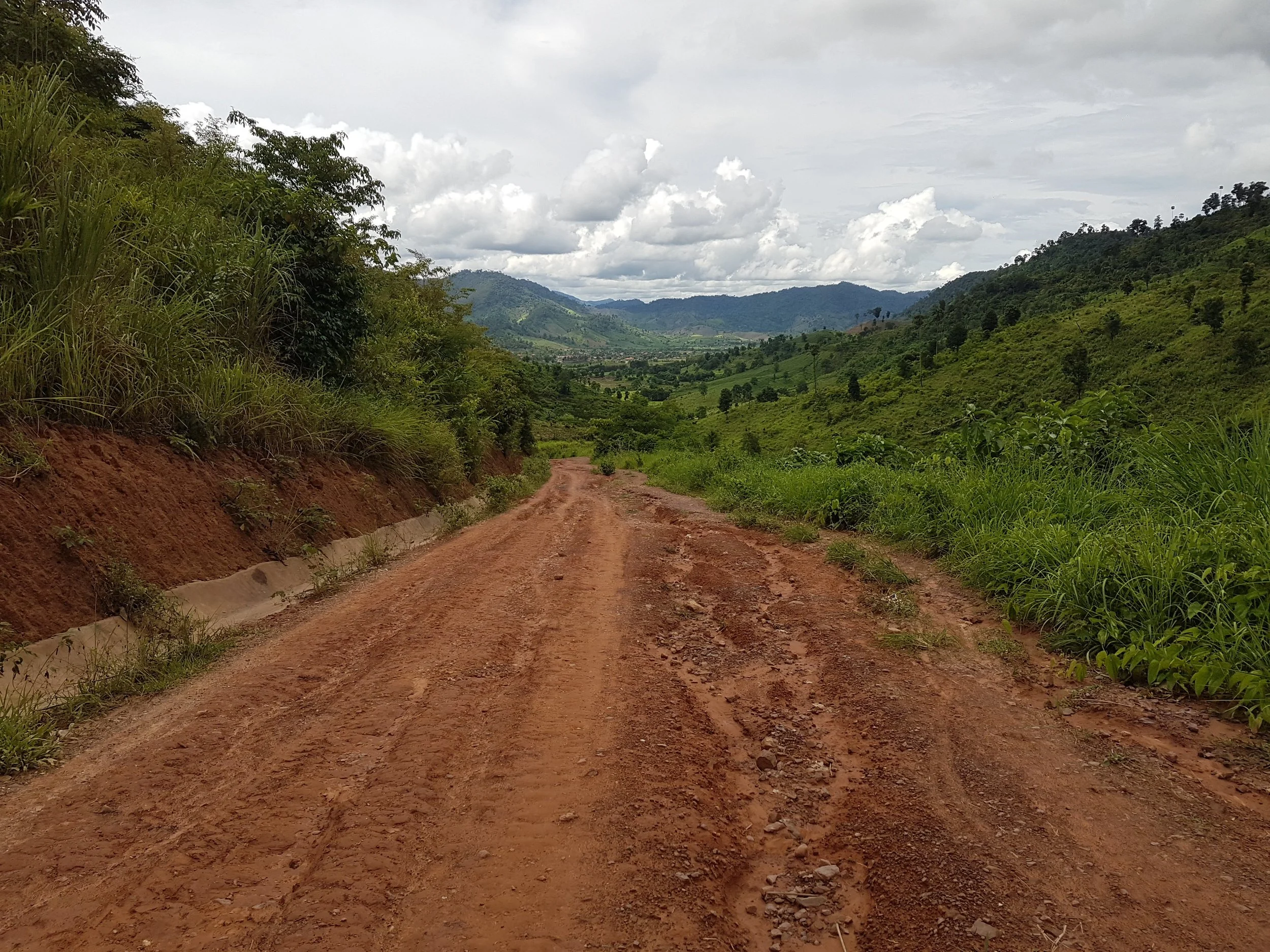 Chemin en terre rouge entouré de verdure avec des montagnes au loin et un ciel nuageux.