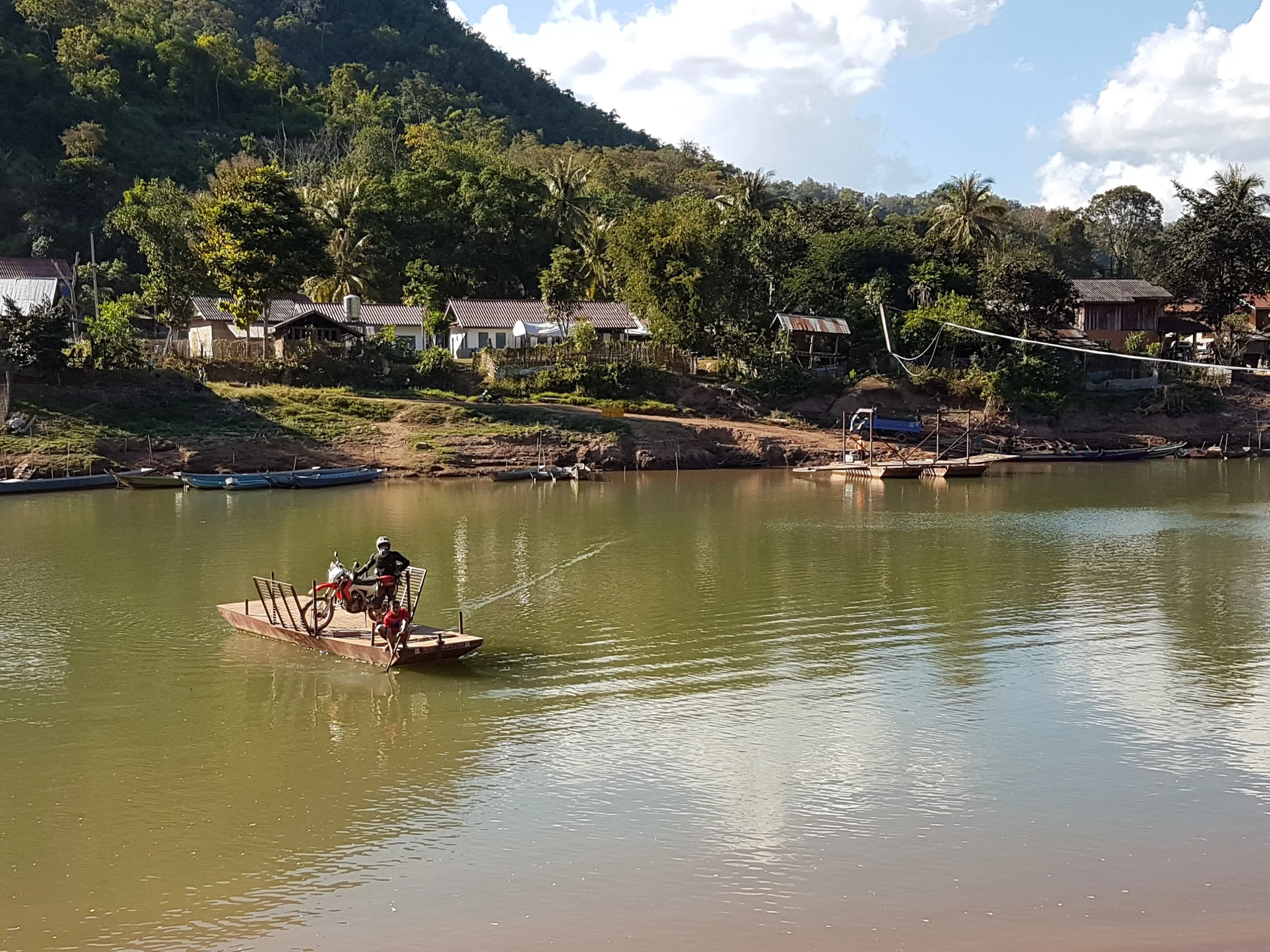 Une scène de rivière avec des petites maisons en arrière-plan entourées d'arbres et de collines, avec un bateau en bateau à moteur et un ponton en bois dans l'eau.