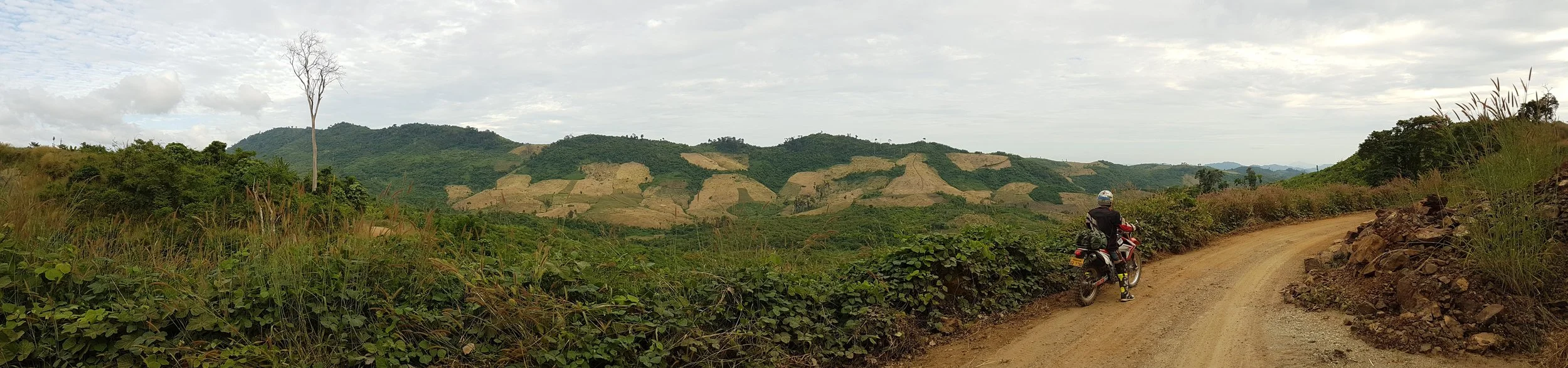 Un motard en tenue noire, casque et gants, assis à côté de sa moto sur un chemin de terre dans un paysage rural avec des collines verdoyantes et des champs en terrasse, sous un ciel nuageux.