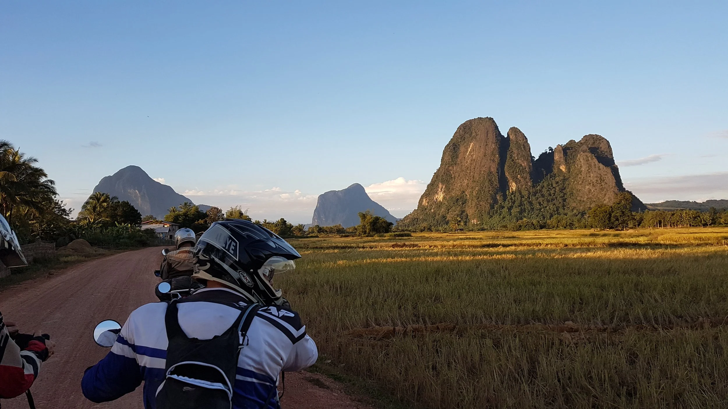 Paysage rural avec des montagnes en arrière-plan, un champ vert à l'avant, des motos garées sur une route en terre, et des arbres au bord de la route.