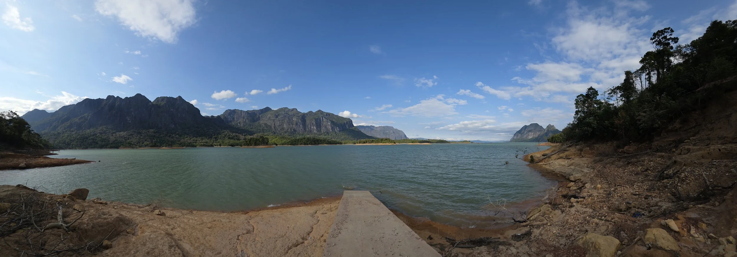 Lac entouré de montagnes escarpées sous un ciel bleu avec quelques nuages, vue panoramique depuis une rive rocheuse.