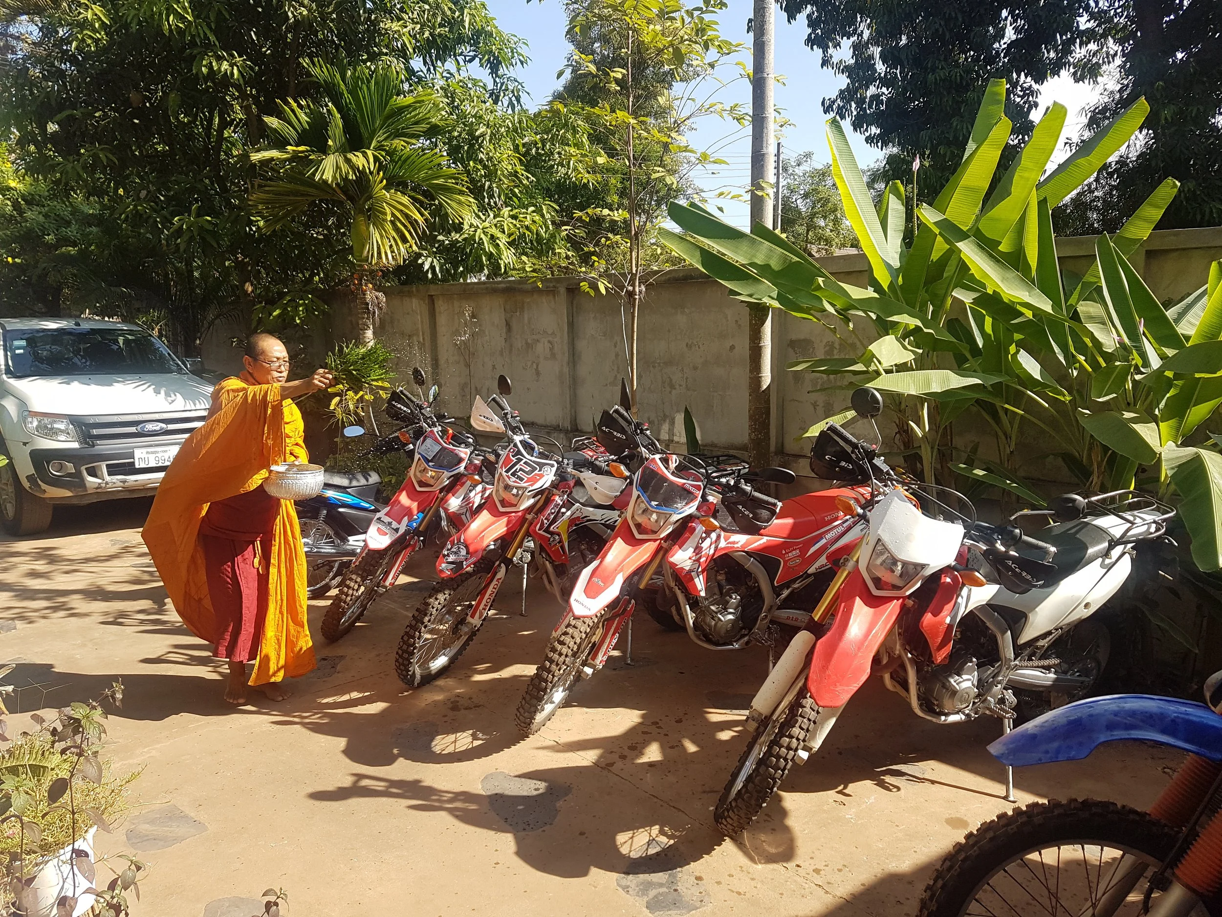 Un homme en vêtements traditionnels oranges effectue une cérémonie dans la cour, entouré de motos et d'une voiture, avec des arbres verts en arrière-plan.