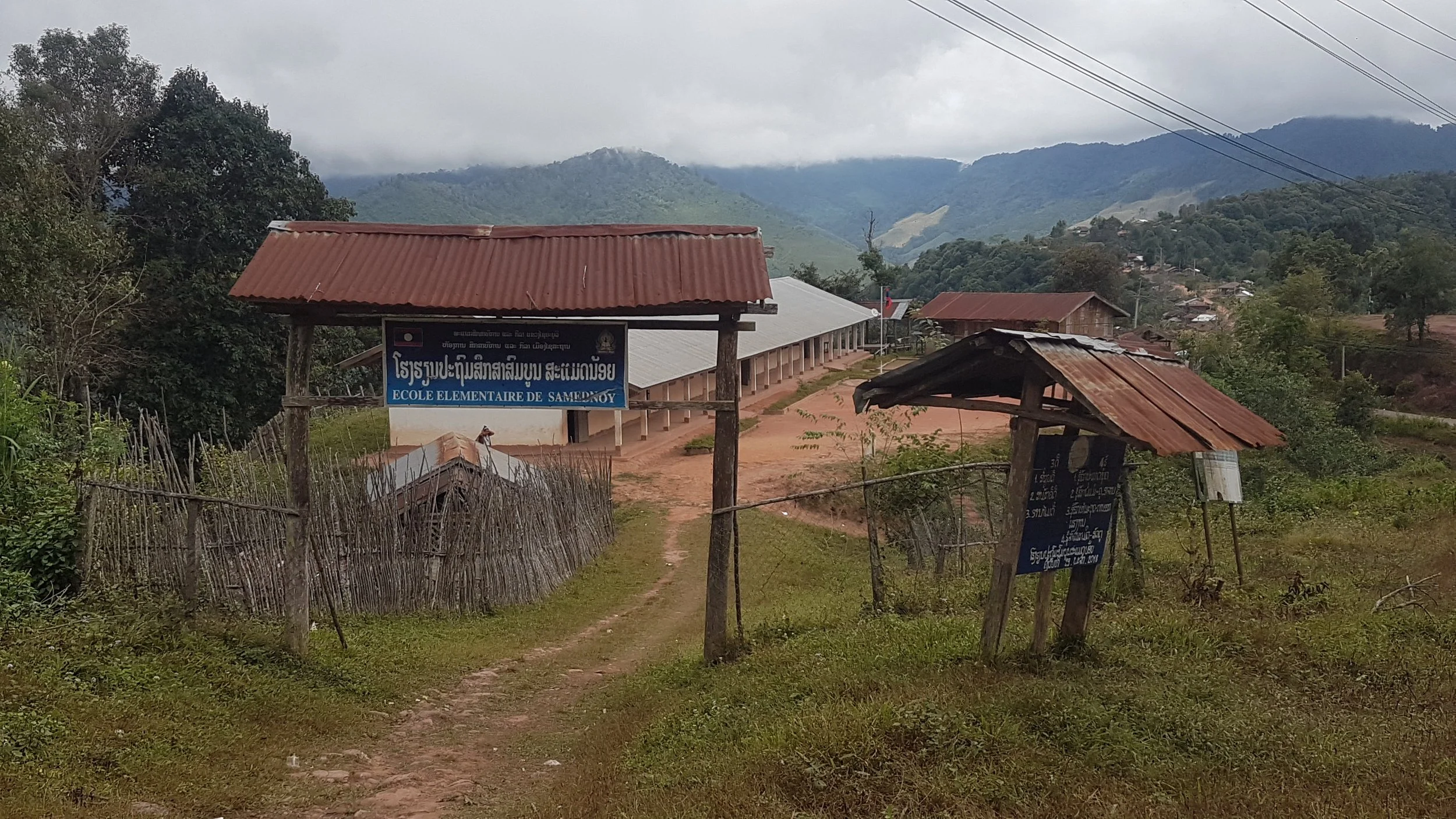 Vue d'une école élémentaire en milieu rural avec des montagnes en arrière-plan, des bâtiments en bois et des panneaux d'information à l'entrée.