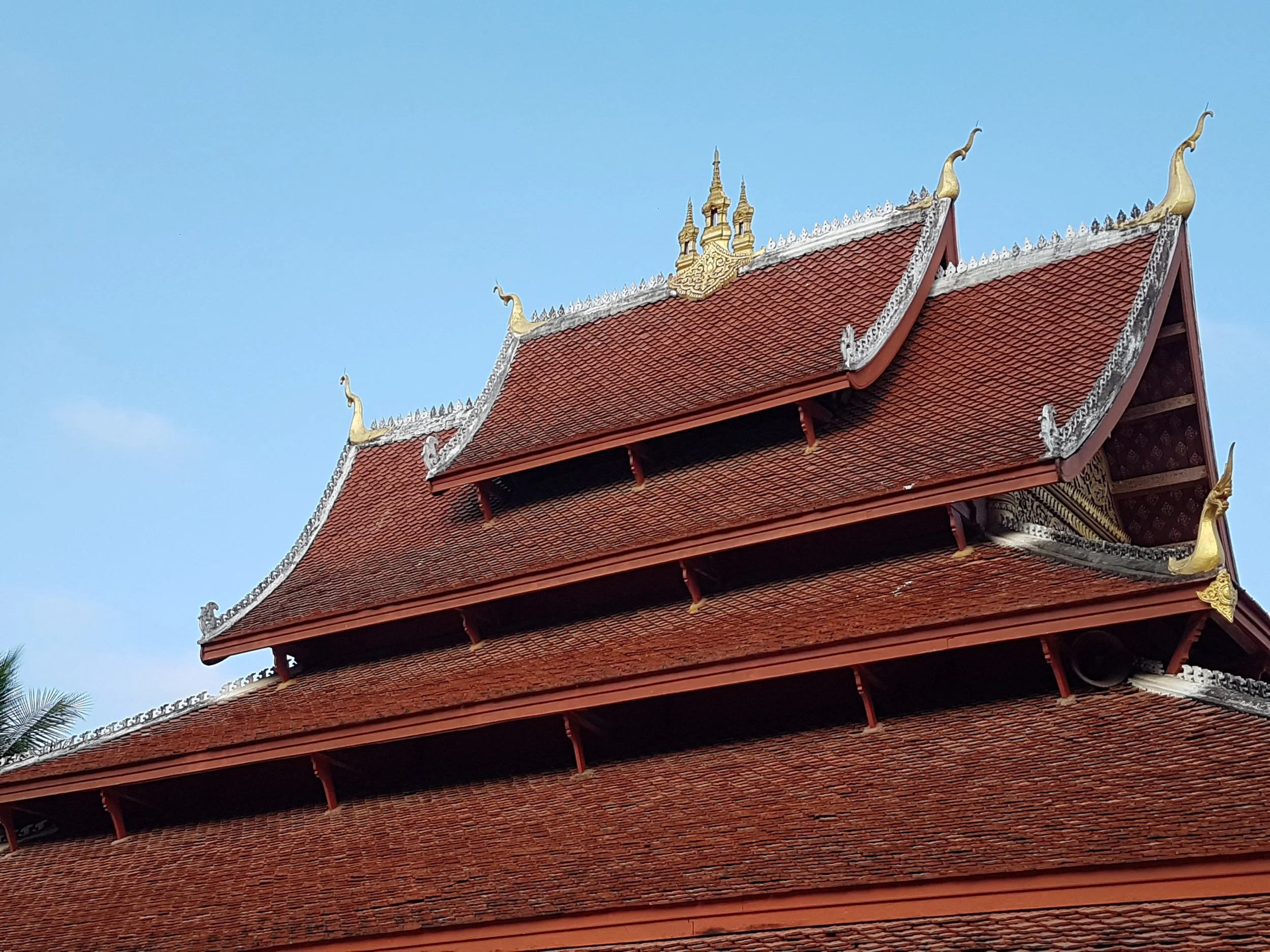Toit d'une structure traditionnelle thaïlandaise avec des détails dorés et des éléments en forme de serpent, sous un ciel bleu clair.