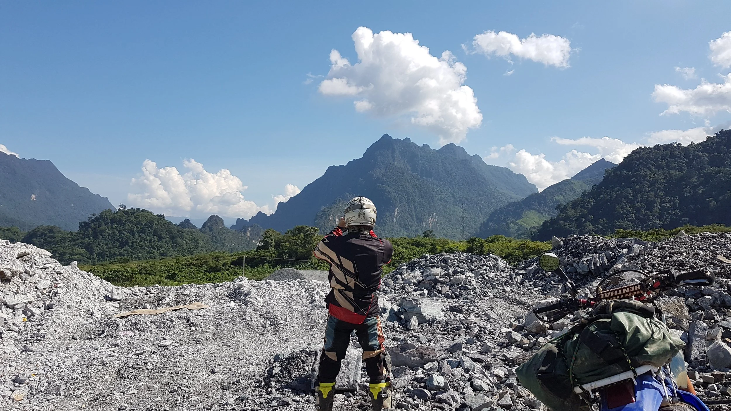 Personne en tenue de moto, portant un casque, regardant un paysage de montagnes verdoyantes sous un ciel bleu avec des nuages blancs, dans un endroit rocailleux.