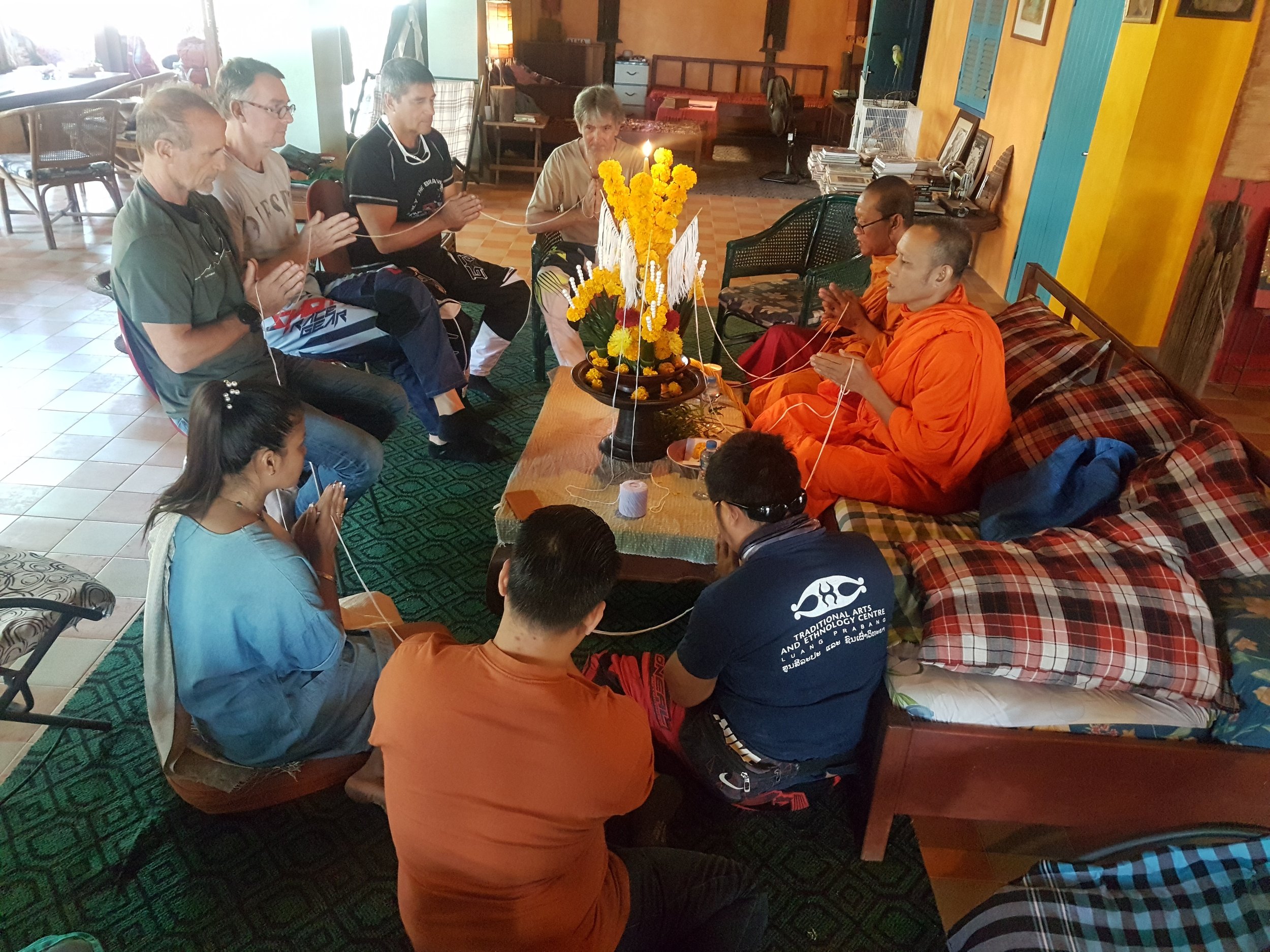 Groupe de personnes assises en cercle, dont deux moines en habits orange, dans une pièce décorée, participant à une cérémonie ou prière devant un arrangement floral avec une bougie.