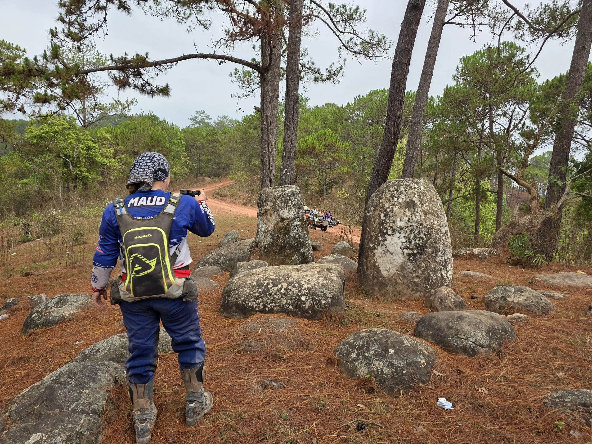 Une personne portant un sac à dos et un casque tourne le dos, marchant sur un sentier forestier avec des rochers et des arbres verts abondants. Au fond, on voit un groupe de personnes en quads ou motos.