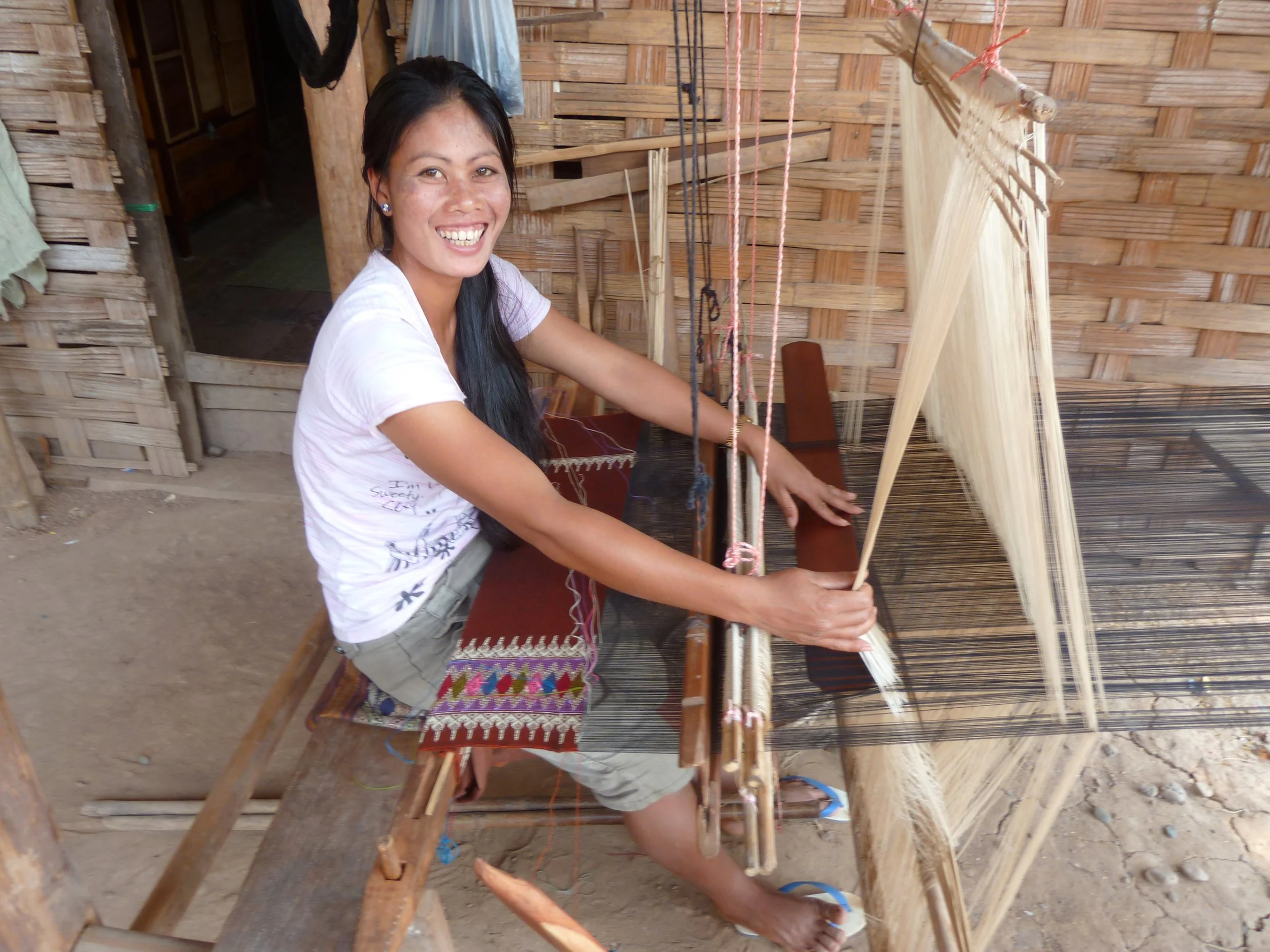 Une femme assise devant un métier à tisser en bois, dans une maison en bambou, souriante.