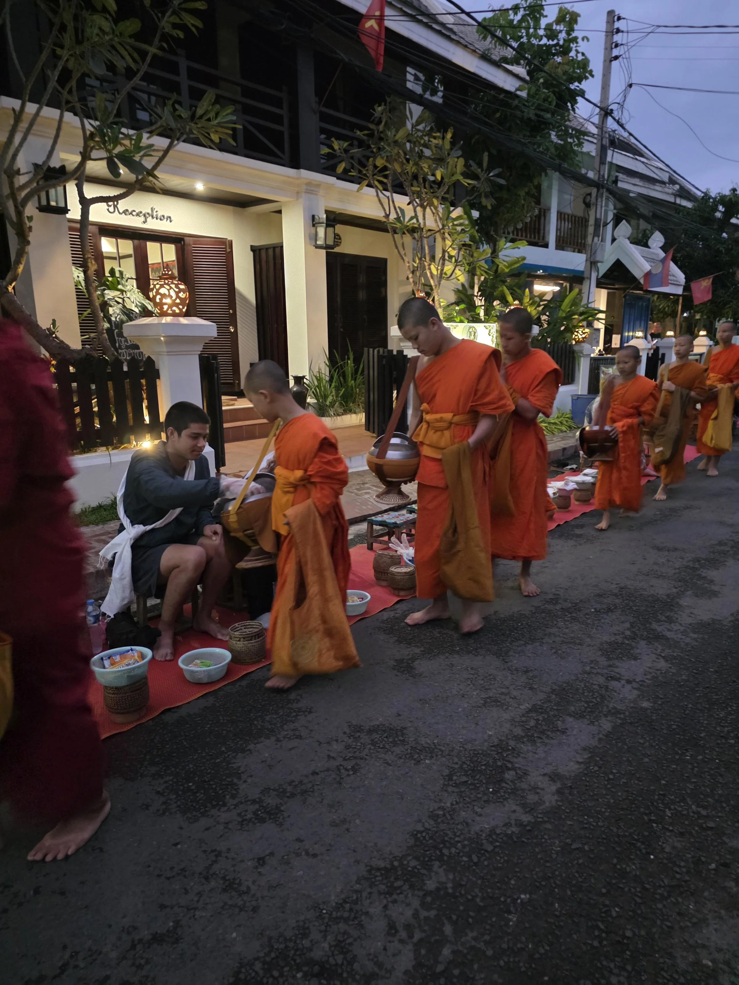 Procession de moines bouddhistes en robes orange, marchant pieds nus dans une rue, avec un homme en tenue en bas à gauche et une maison en arrière-plan.
