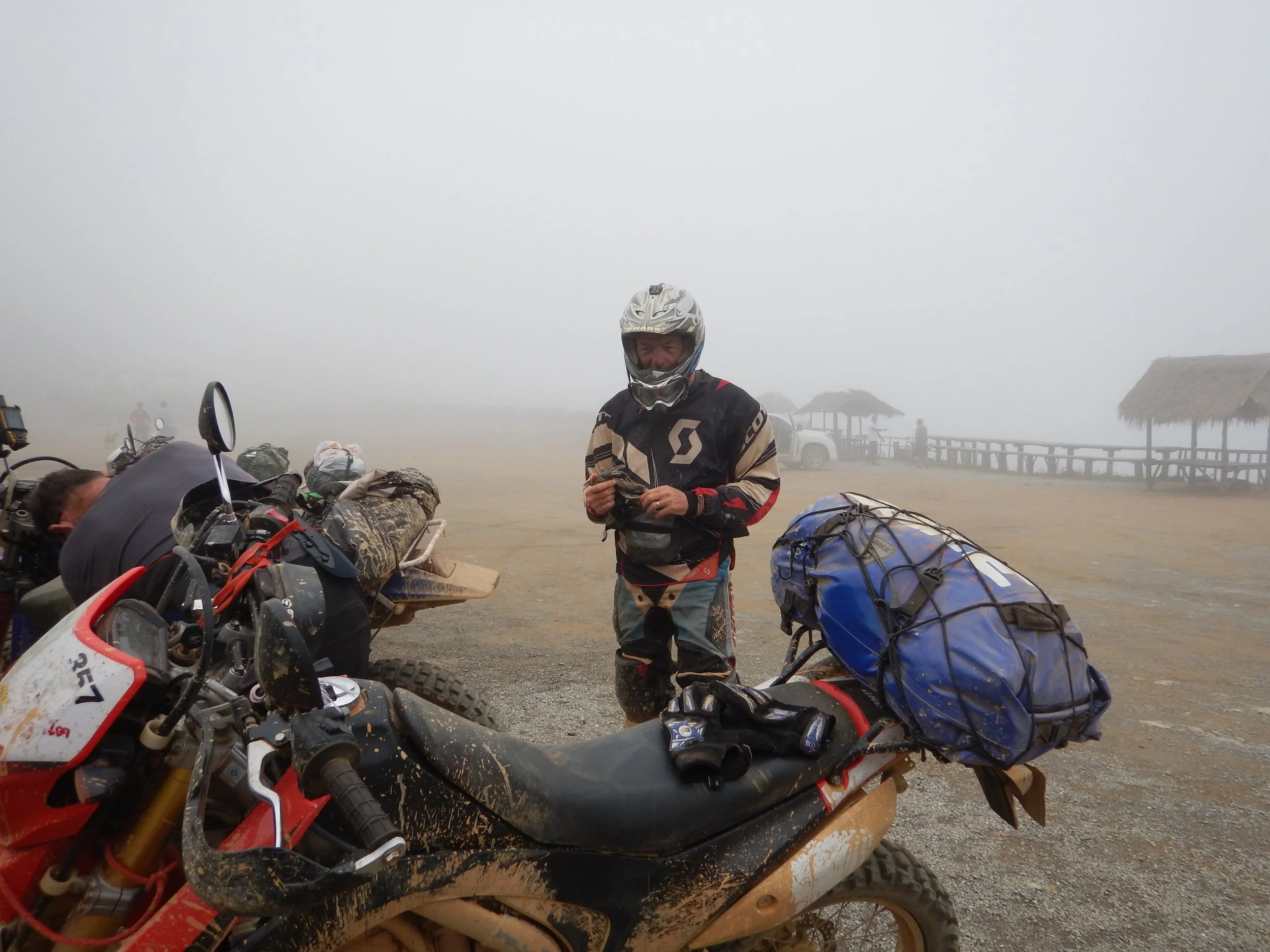 Une personne portant un casque de motocross, debout parmi des motos tout-terrain couvertes de boue, dans un environnement brumeux ou poussiéreux avec des structures en bambou en arrière-plan. Montagne de Kasi en direction de Luang Prabang Laos.