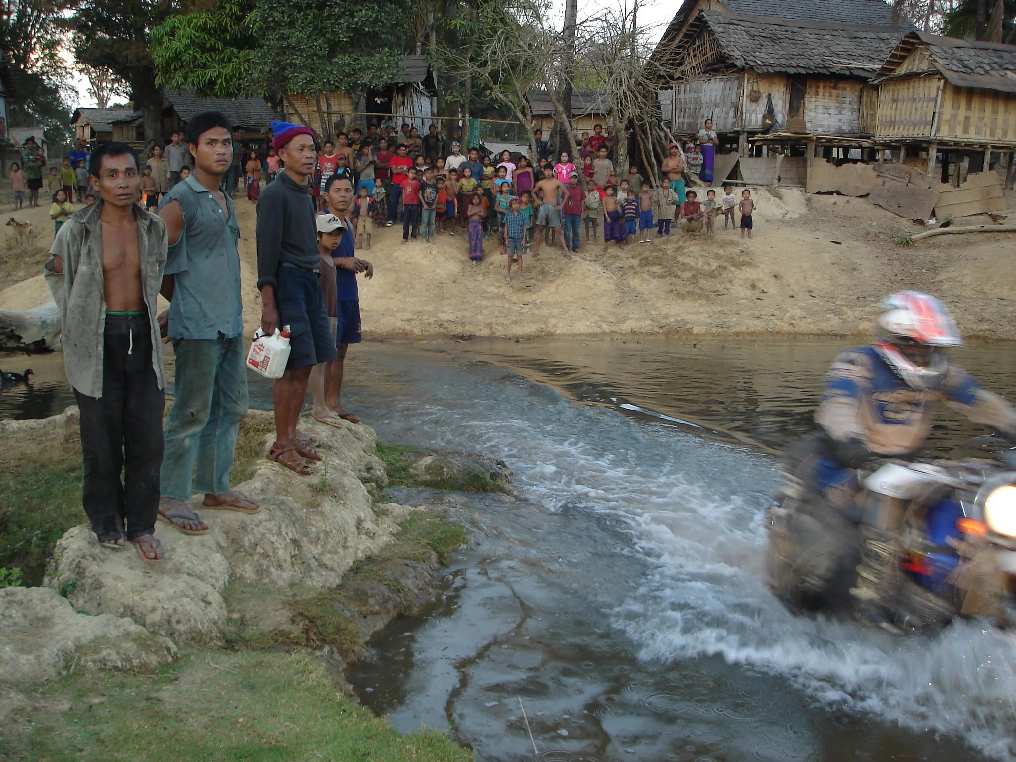 Un groupe d'enfants et d'adultes debout sur une berge en terre près d'une rivière ou d'un plan d'eau. Certaines personnes regardent l'eau, tandis qu'une personne à moto passe rapidement dans l'eau, créant une éclaboussure. Les habitations en bois sont visibles en arrière-plan, avec des arbres environnants