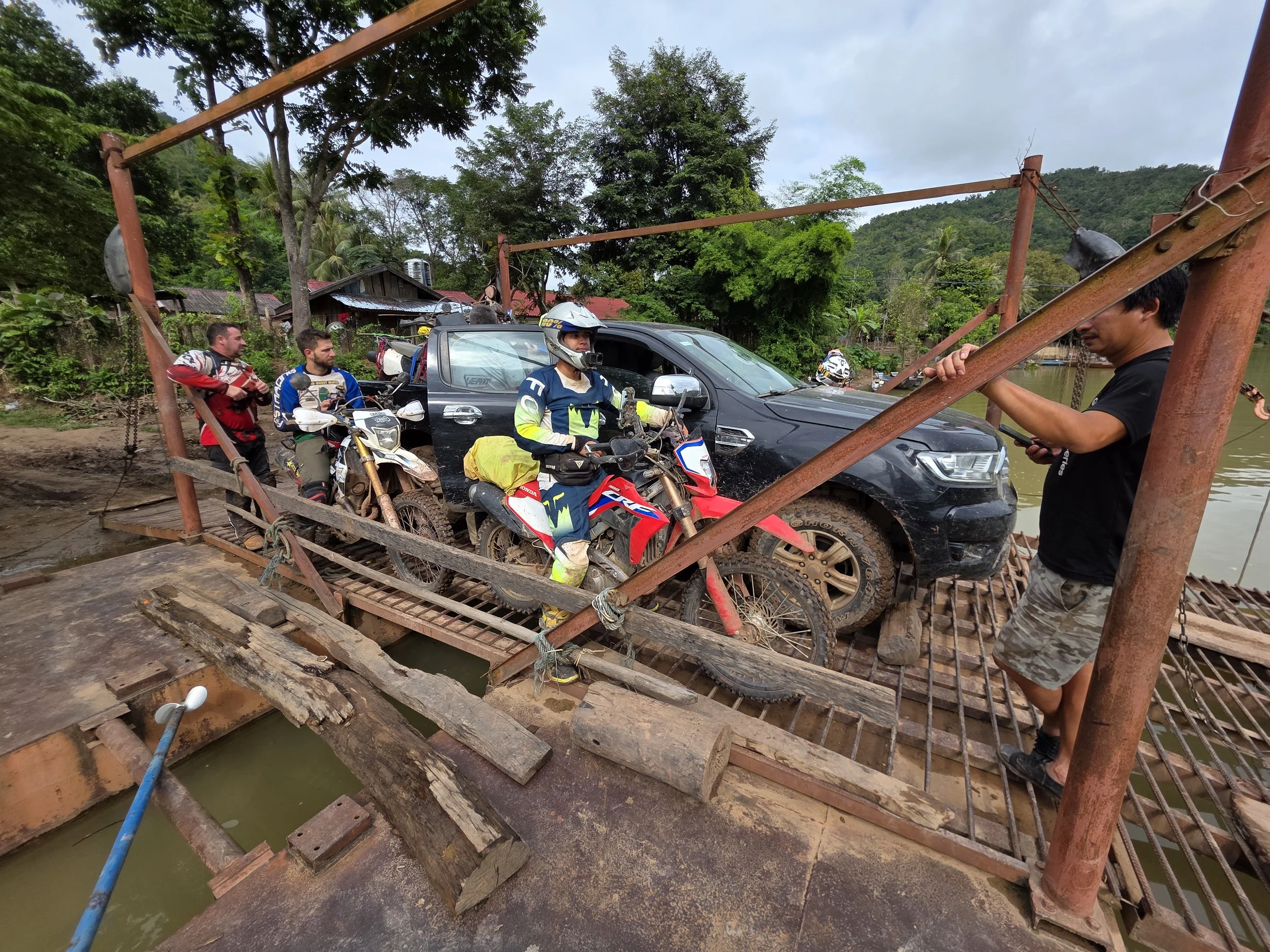 Groupe de personnes, dont certains motocyclistes portant des casques, attendant sur un pont en bois fragile au-dessus de l'eau, entouré de végétation dense.