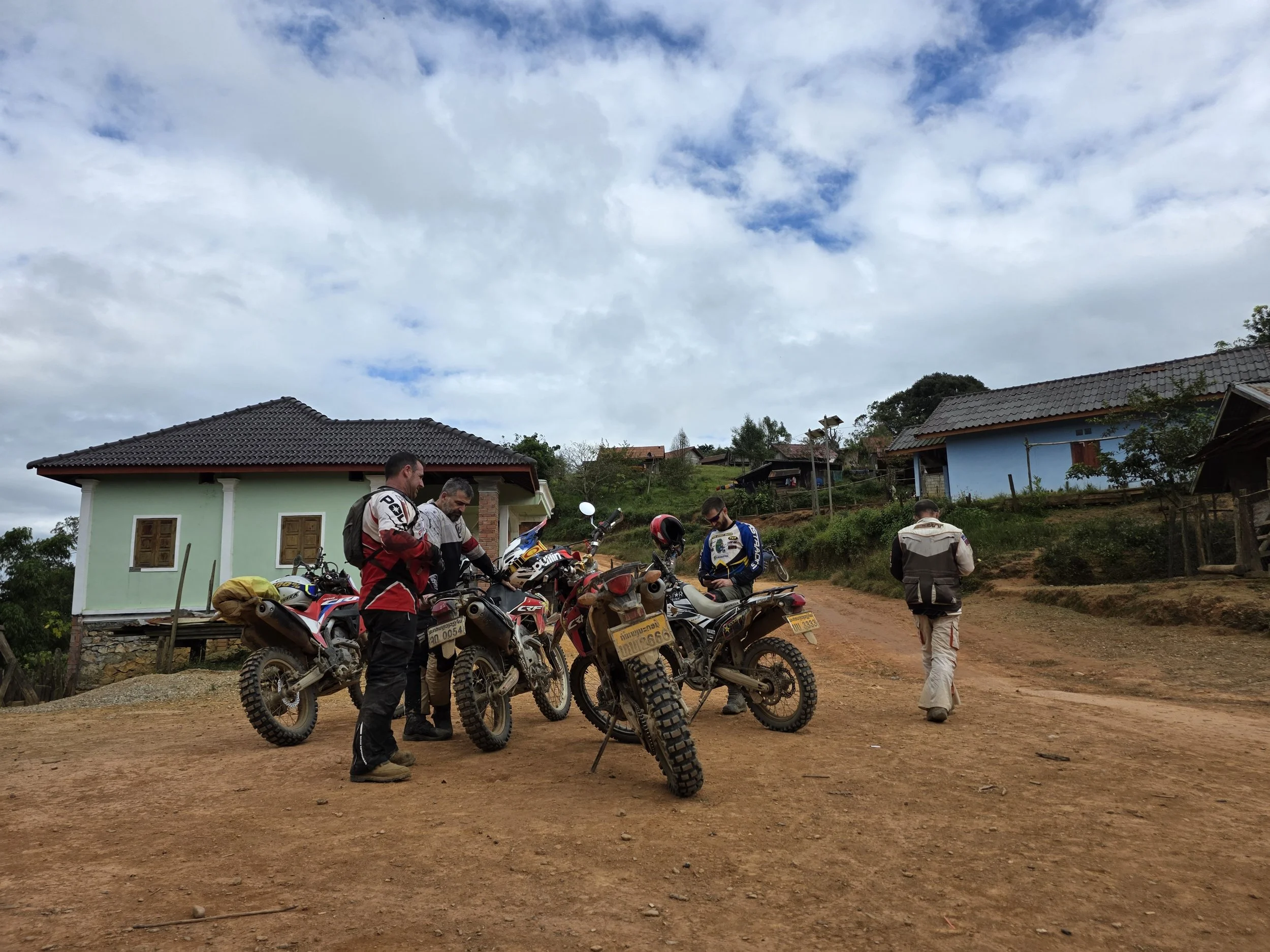 Groupe de motards en tenue de moto, arrivant ou préparant leur sortie, avec plusieurs motos tout-terrain, dans un village rural avec des maisons colorées et un ciel nuageux.