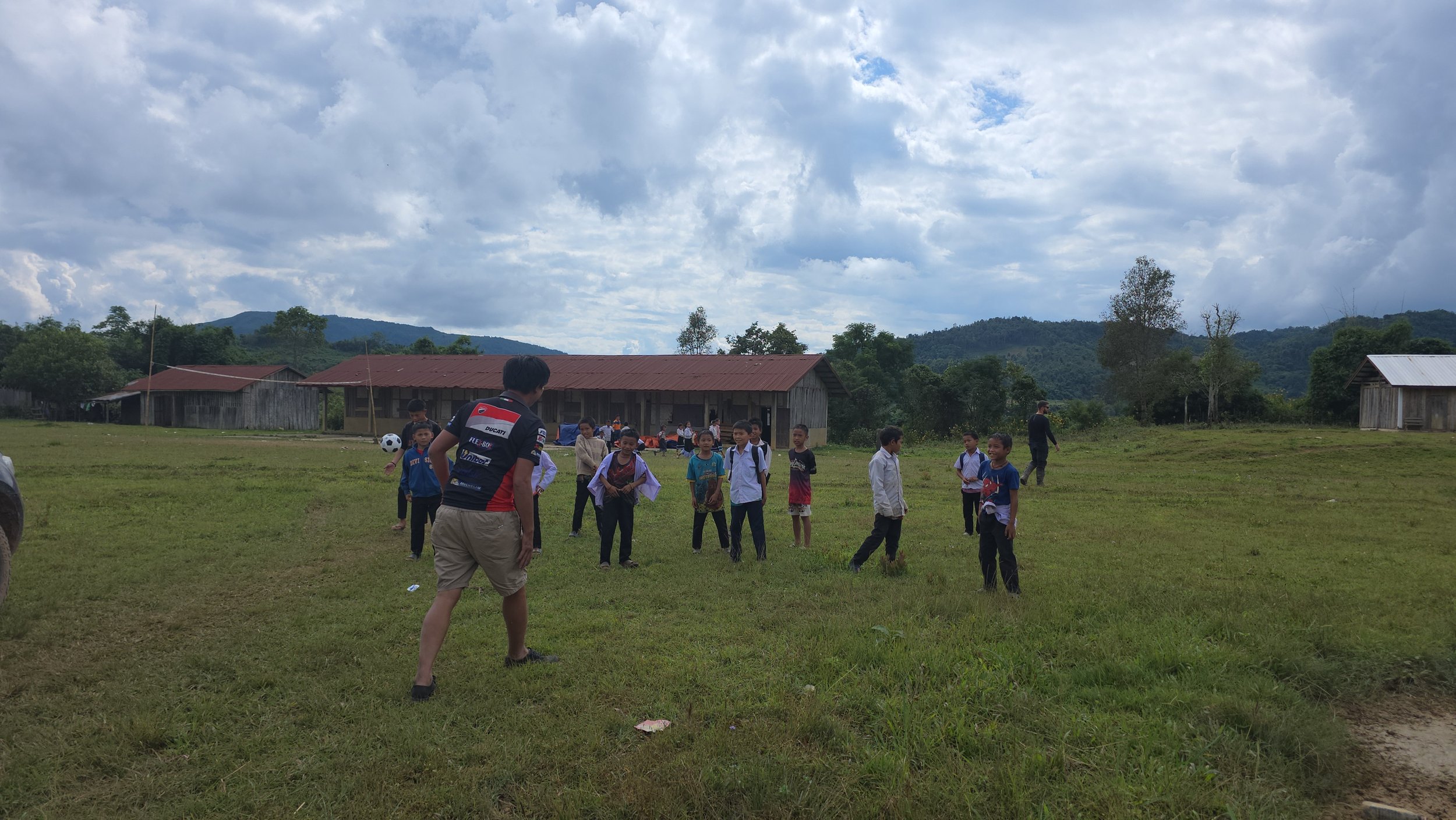 Des enfants jouent au football dans un champ en milieu rural, avec des maisons en bois et des montagnes en arrière-plan, sous un ciel nuageux.
