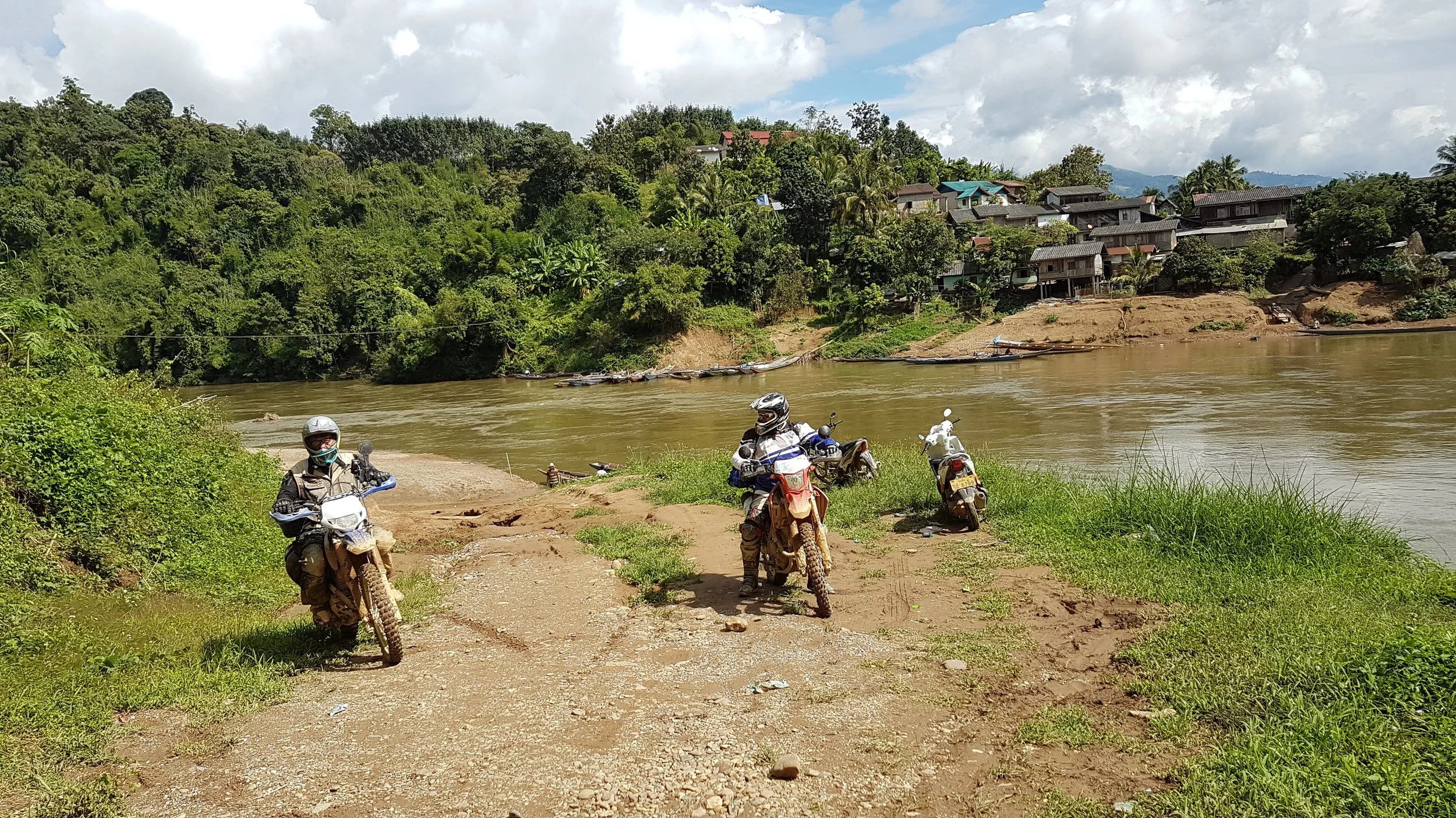 Trois motards en équipement de protection sur un chemin près d'une rivière, avec une ville en hauteur entourée de végétation luxuriante sous un ciel partiellement nuageux.