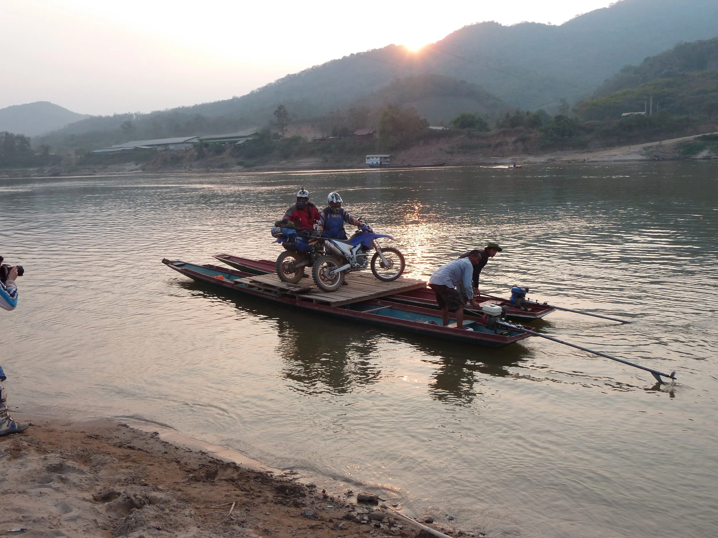 Les gens chargent une moto sur une plateforme flottante sur une rivière, avec un paysage de montagnes et un coucher de soleil en arrière-plan.
