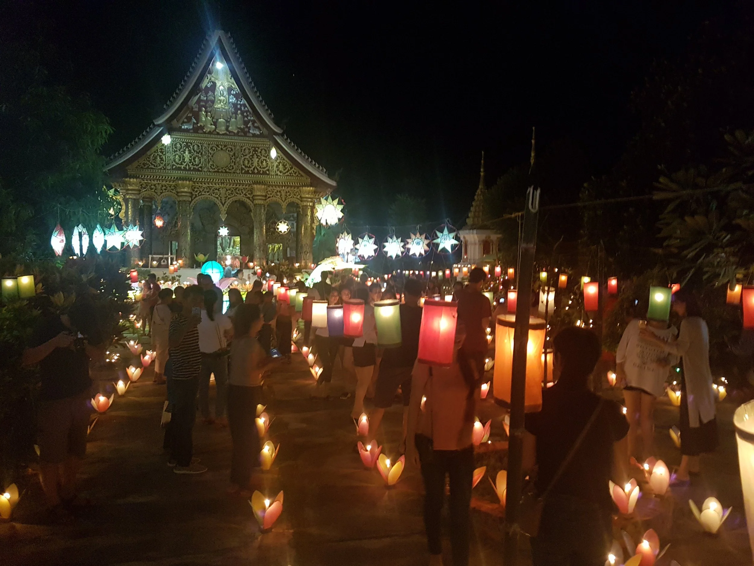 Une scène de nuit avec une structure en bois richement décorée et illuminée, entourée de lanternes colorées suspendues ou posées au sol, avec des personnes assises ou se promenant, dans un événement culturel ou festival.