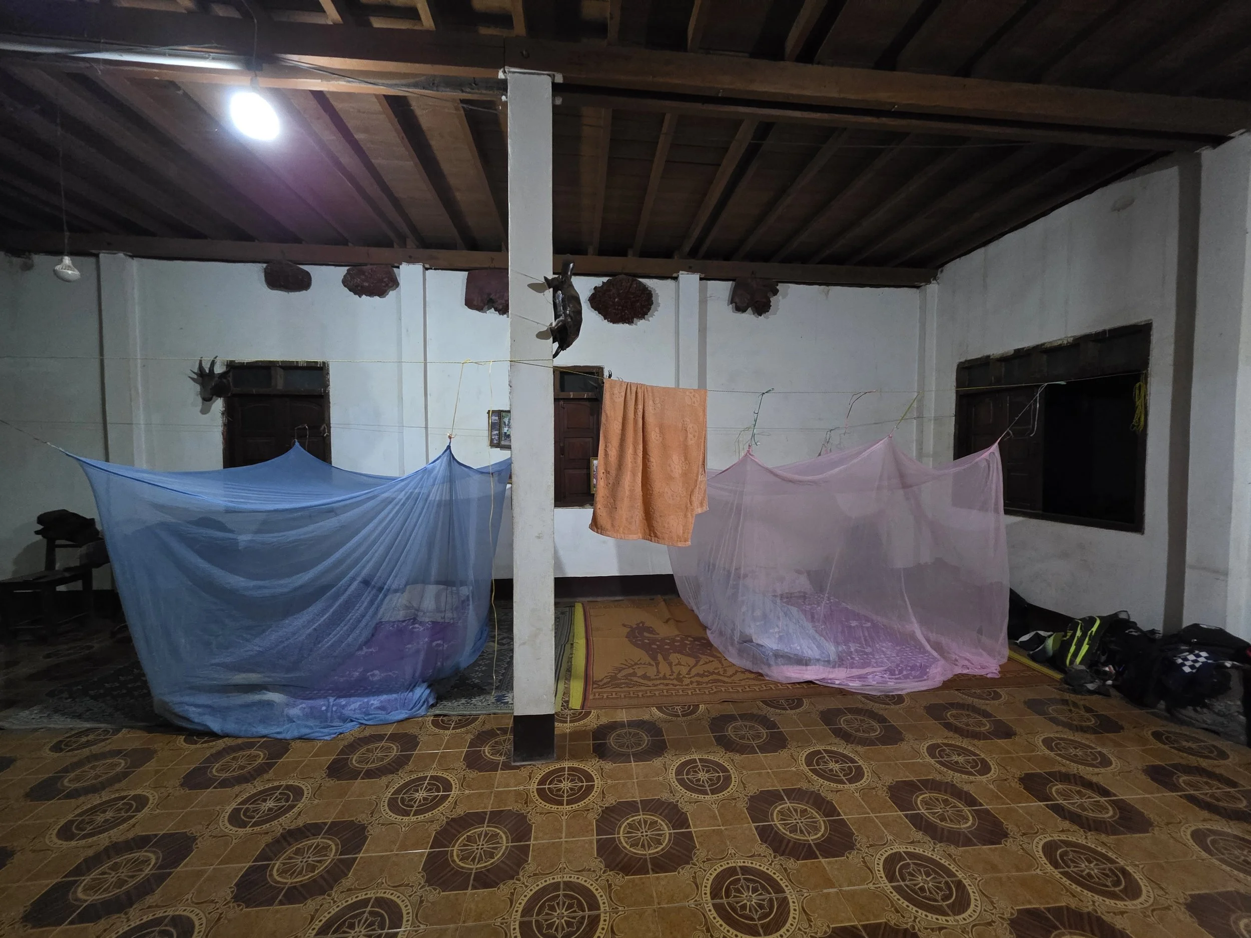 Intérieur d'une pièce avec plafond en bois, murs blancs, et plusieurs draps suspendus, probablement pour sécher. Le sol est en carrelage avec un motif circulaire. On voit des chaussures et des sacs sur le côté droit.