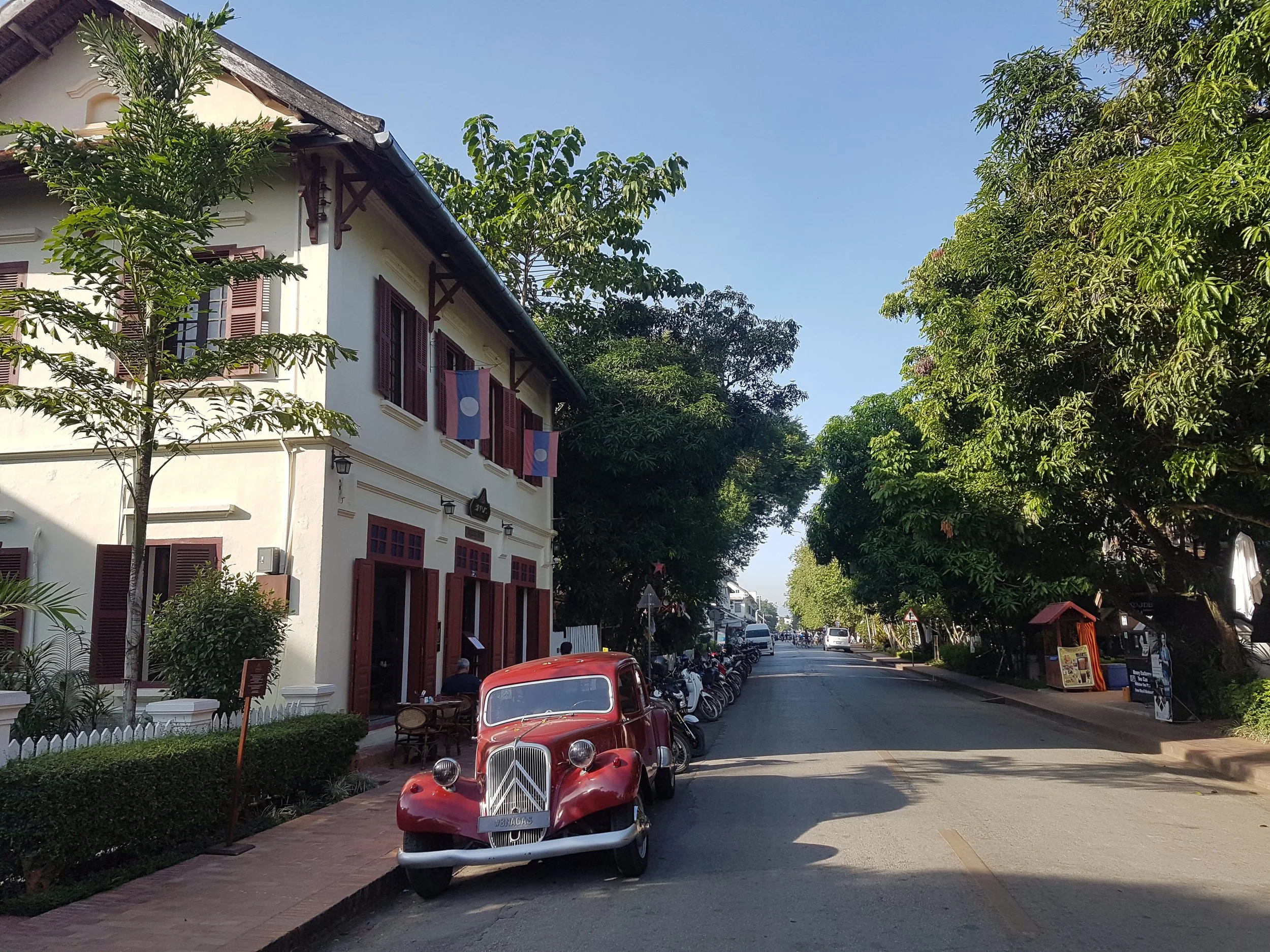 Une rue bordée d'arbres avec une maison blanche à fenêtres à volets marron, des voitures anciennes rouges garées sur le trottoir et des drapeaux du Laos accrochés à la façade.