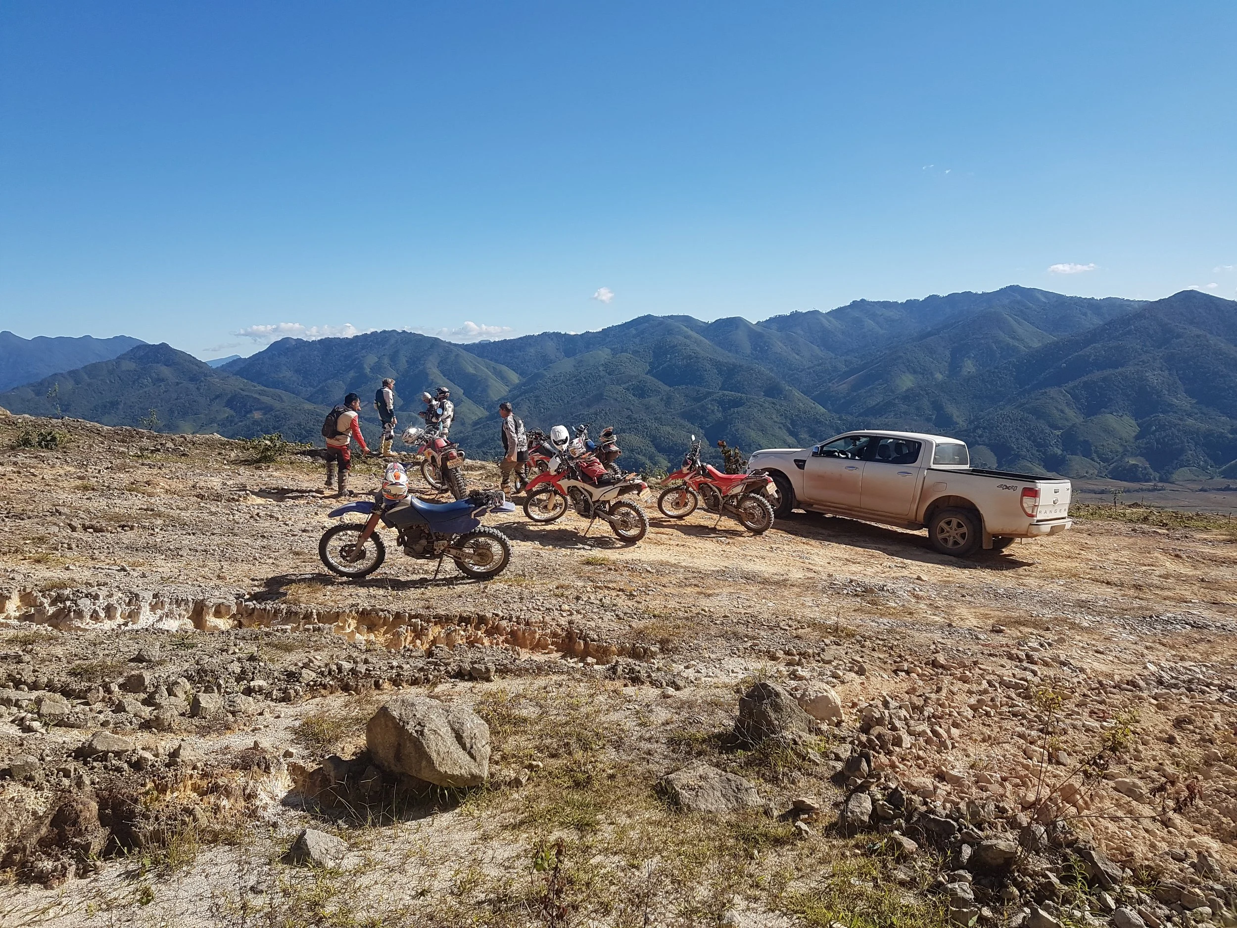 Groupe de motards avec casques et motos tout-terrain, certains discutant ou préparant leurs véhicules, avec un véhicule pickup blanc garé à côté, dans un paysage de montagnes et ciel clair. Au Laos.
