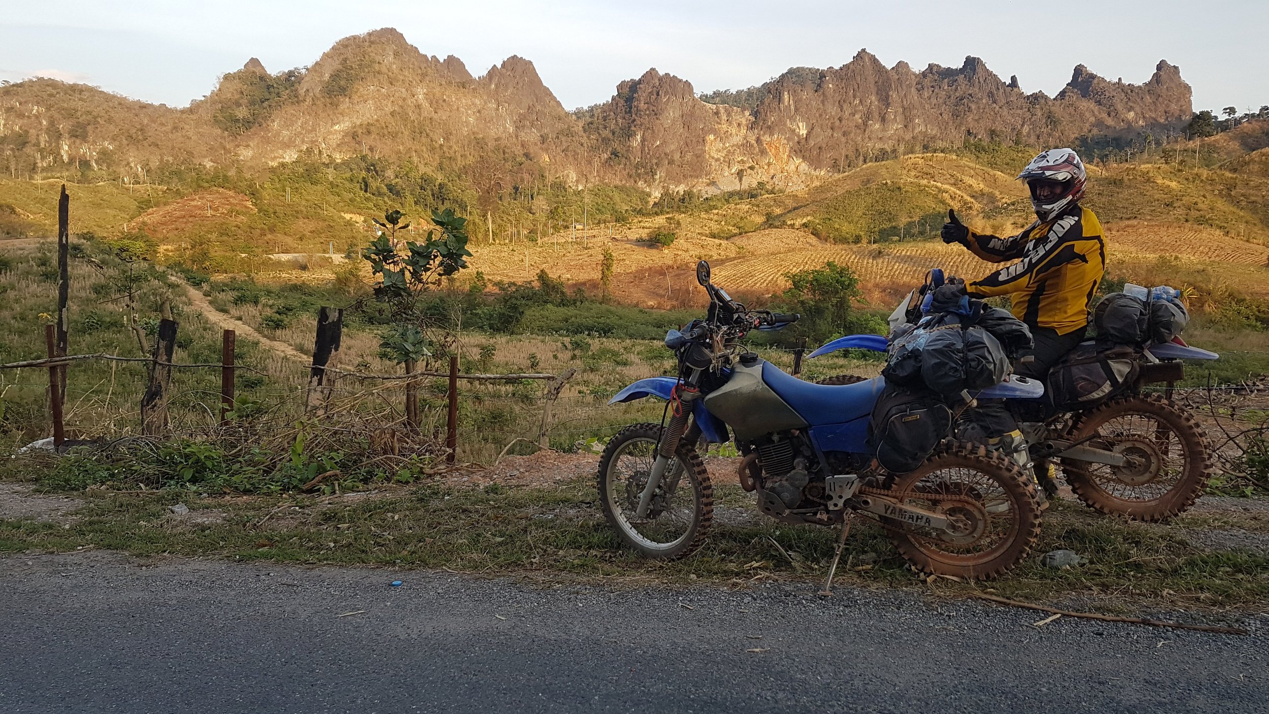 Motard en équipement de protection donnant un signe de pouce en l'air, à côté de sa moto endommagée, avec un paysage de montagnes et de paysages agricoles en arrière-plan.