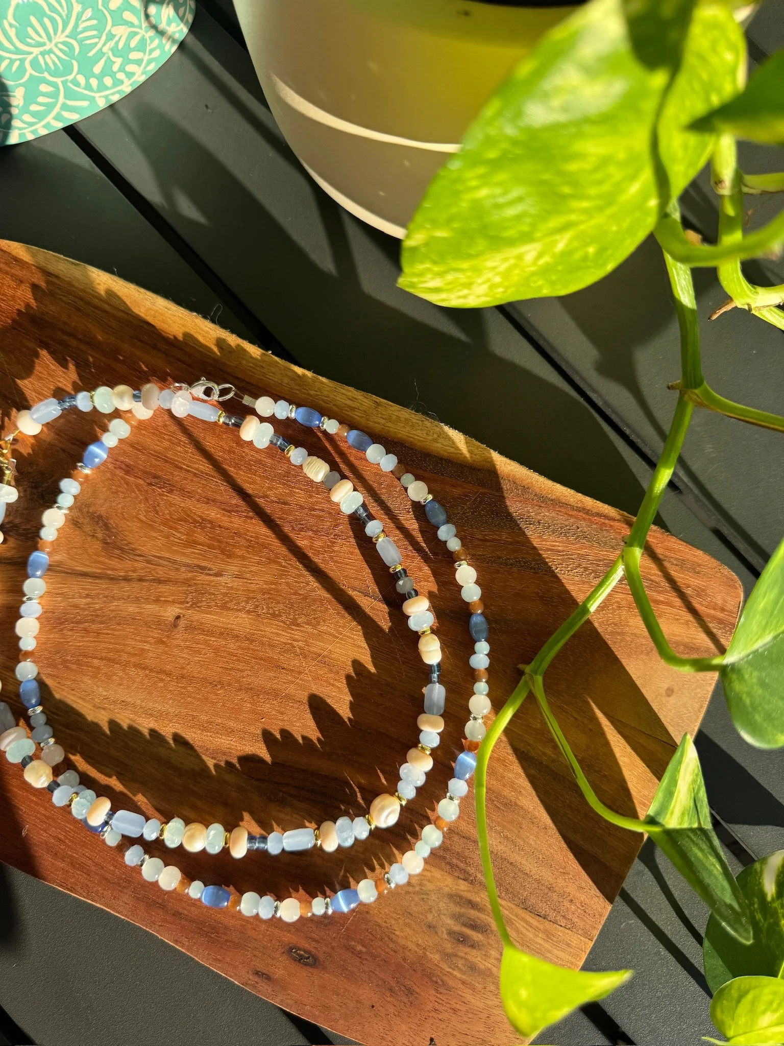 Close-up view of a beaded necklace with white, blue, and clear beads resting on a wooden surface, with green leafy plants and potted plants in the background.