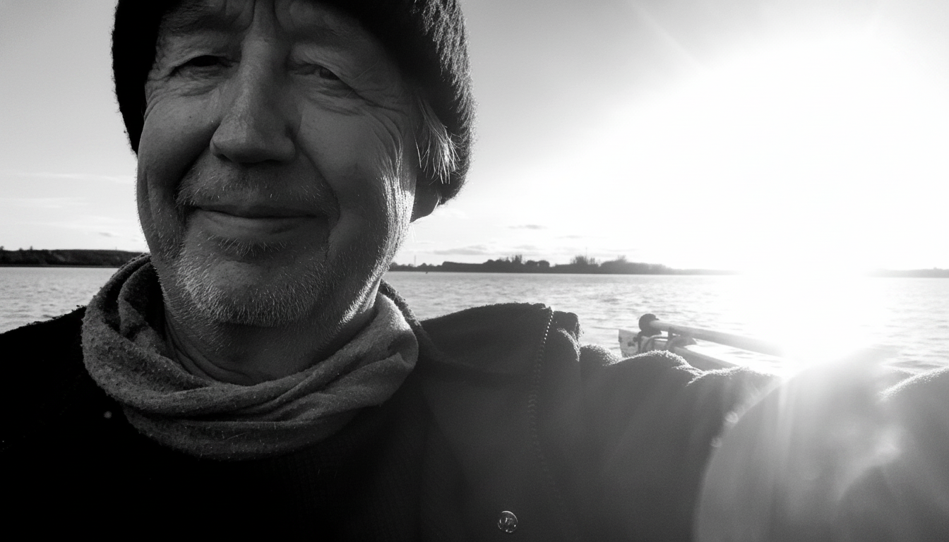Ein älterer Mann macht ein Selfie im Freien bei Sonnenuntergang am Wasser, in Schwarz-Weiß.