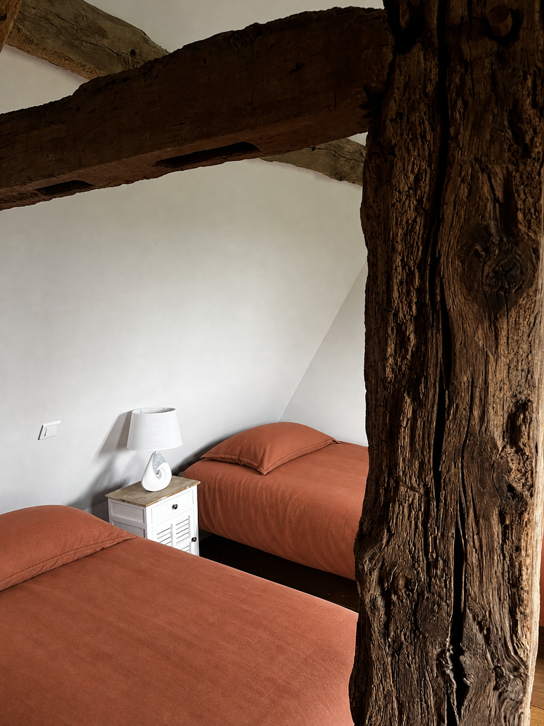Deux lits simples avec des draps et oreillers en couleur terre cuite, une table de chevet blanche avec une lampe moderne à abat-jour blanc, dans une chambre charmante avec mur blanc et poutres en bois apparentes, chambre pour retraite en Normandie