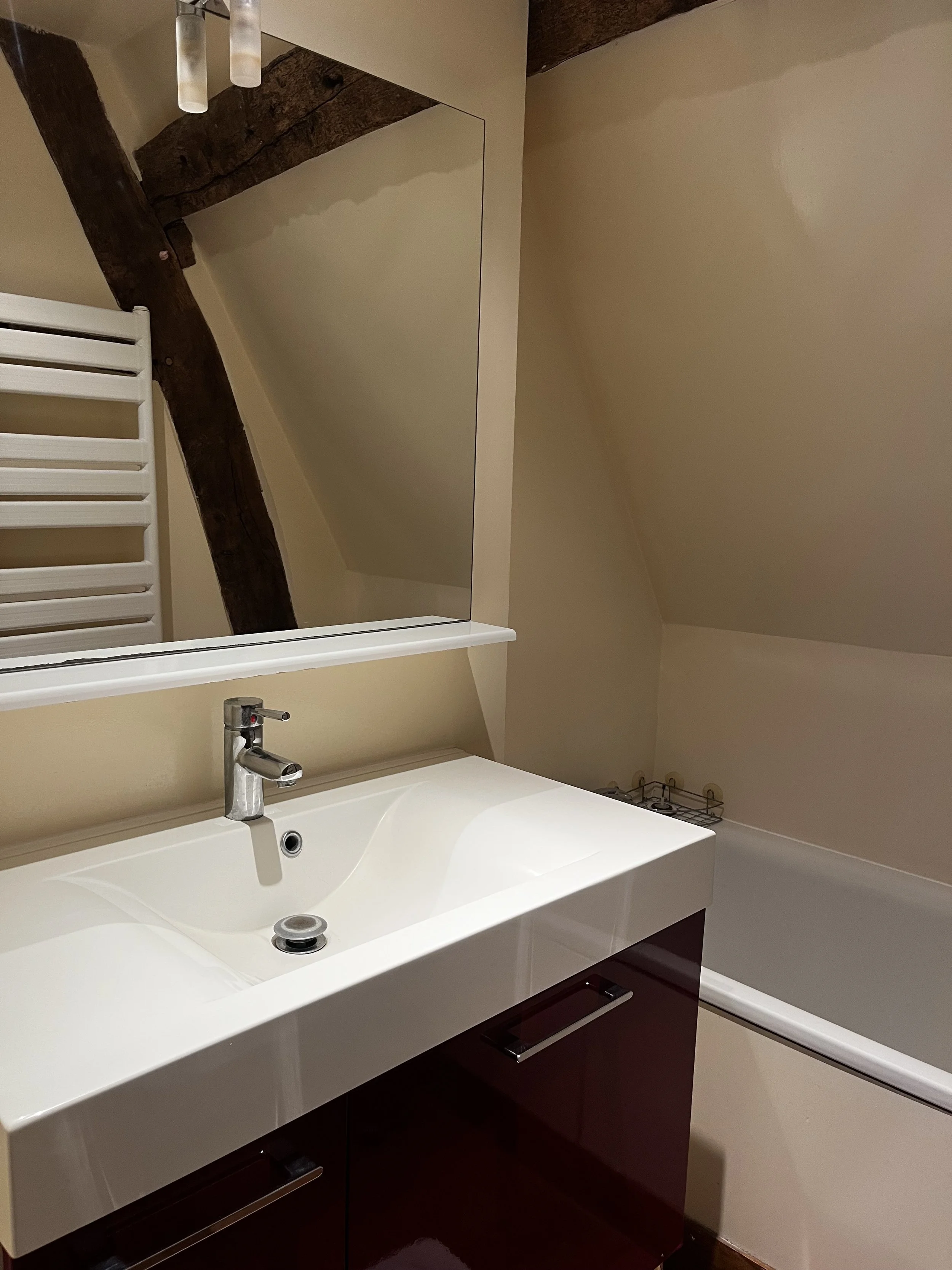 Un lavabo blanc avec un robinet en métal argenté, un miroir au-dessus, un radiateur blanc en forme de grille, et des murs peints en beige, dans une salle de bain, dans un gite pour une retraite en normandie 